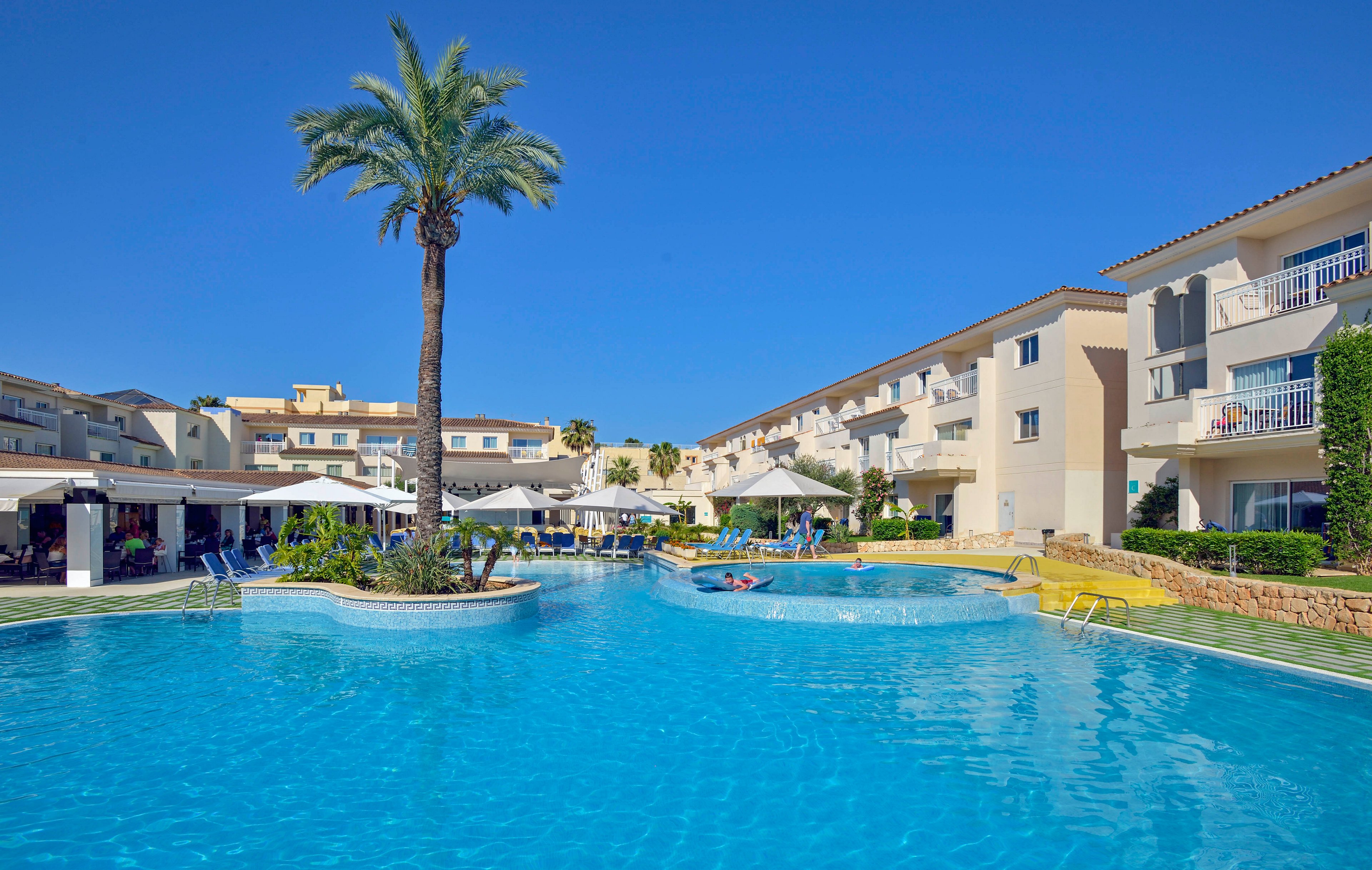 A large outdoor swimming pool is the central feature of this sunny resort scene. The pool is surrounded by modern hotel buildings, sun loungers, and umbrellas, with a tall palm tree adding a tropical touch. Guests are relaxing in and around the pool, enjoying the clear blue sky and tranquil atmosphere. The setting suggests a vacation or leisure environment, with bright, inviting visuals.