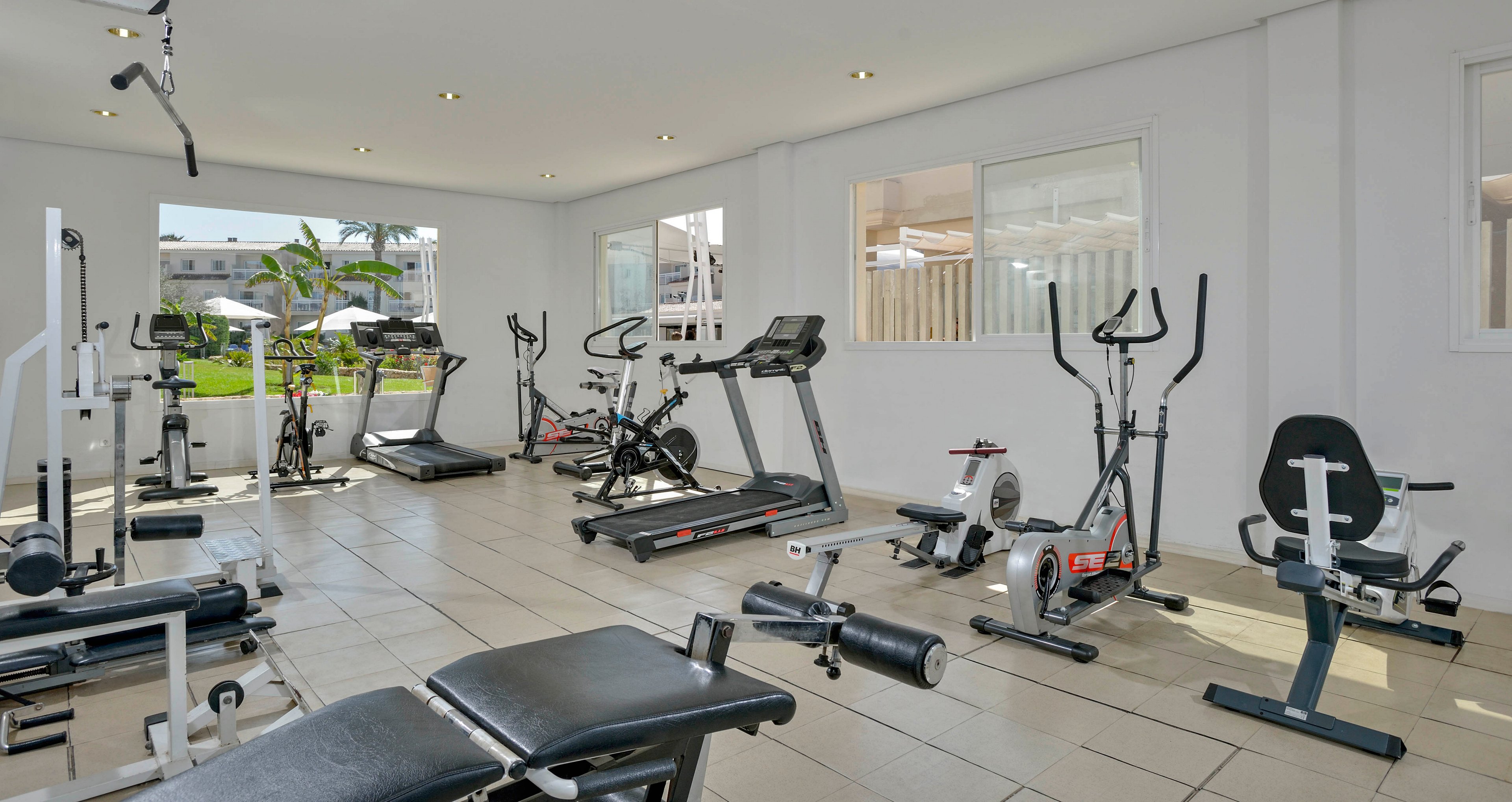 A bright, spacious gym features a variety of exercise machines including treadmills, stationary bikes, ellipticals, and weight benches. The room is well-lit with large windows allowing natural light and views of greenery outside. The clean, minimalist interior uses neutral tones, creating an inviting atmosphere for workouts. No people are present, emphasizing the equipment and open space.