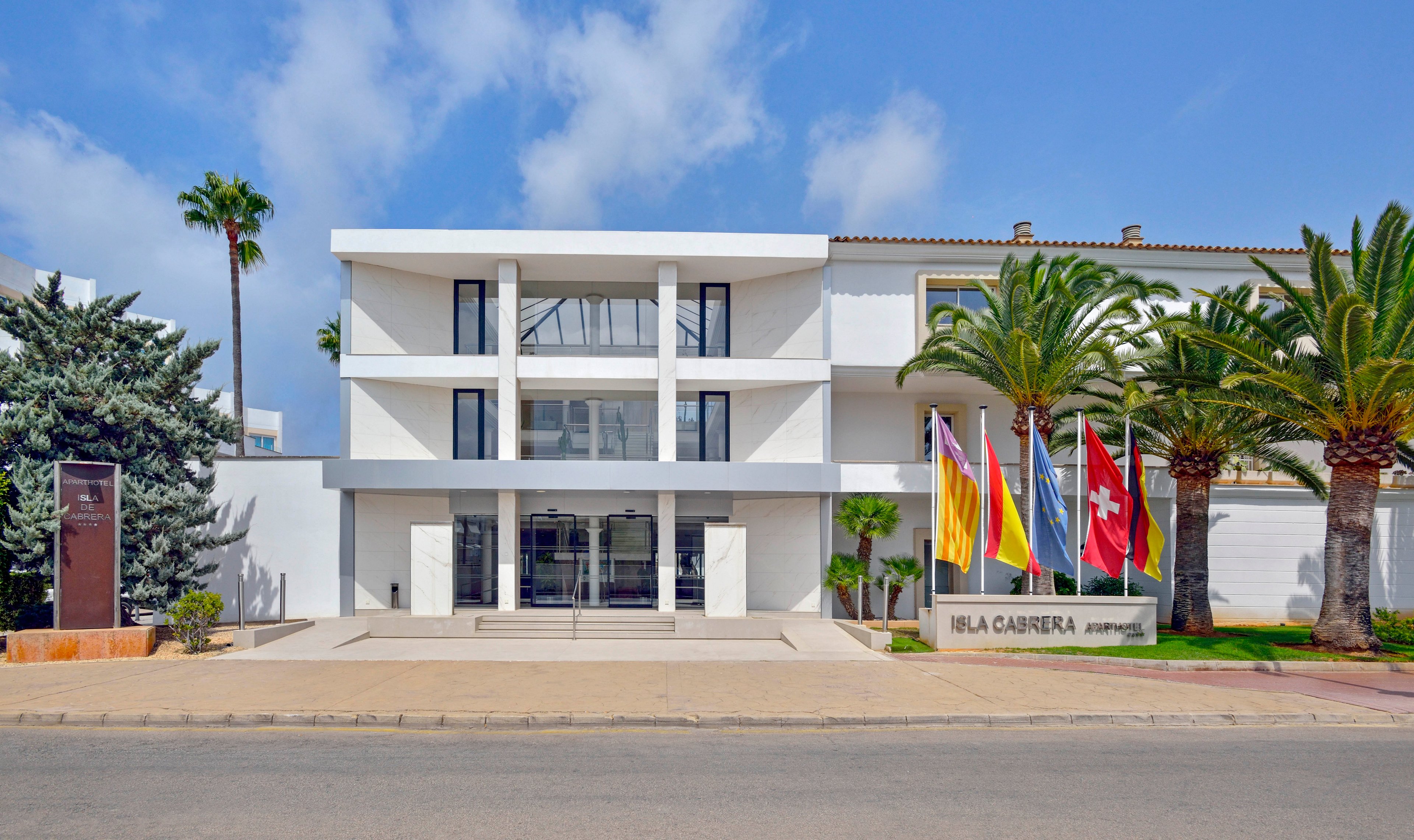 A contemporary white hotel building features a symmetrical facade and large glass entrance. Several national and regional flags are prominently displayed at the front, adding color to the scene. Palm trees and manicured landscaping frame the entrance, while a clear blue sky enhances the inviting atmosphere. The sign near the entrance reads 'Isla Canela'.