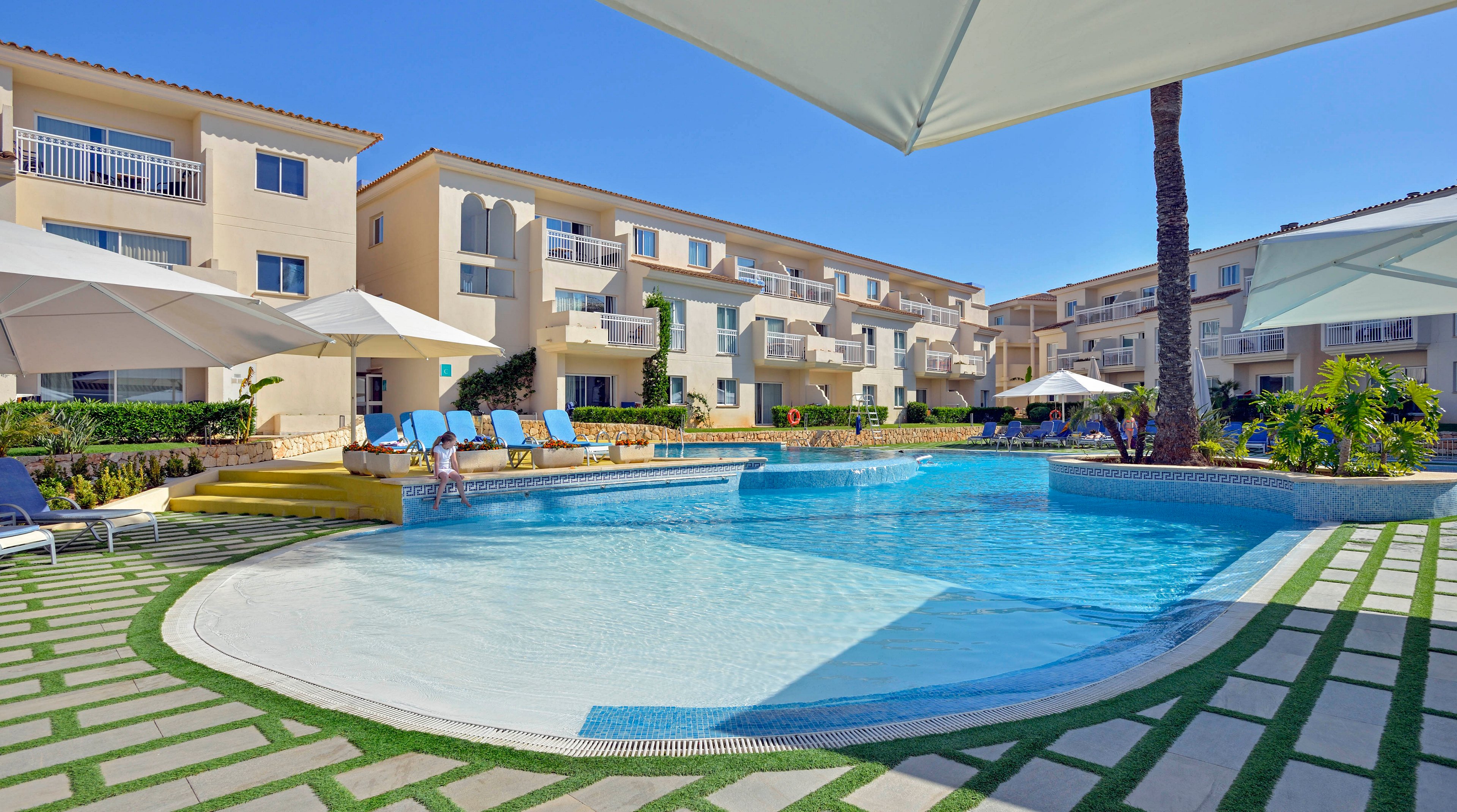 A bright outdoor swimming pool is surrounded by modern resort buildings under a clear blue sky. Several lounge chairs and large white umbrellas provide shaded relaxation areas around the pool. The scene features a clean, inviting atmosphere with green landscaping and tiled walkways, evoking a vacation or leisure setting.