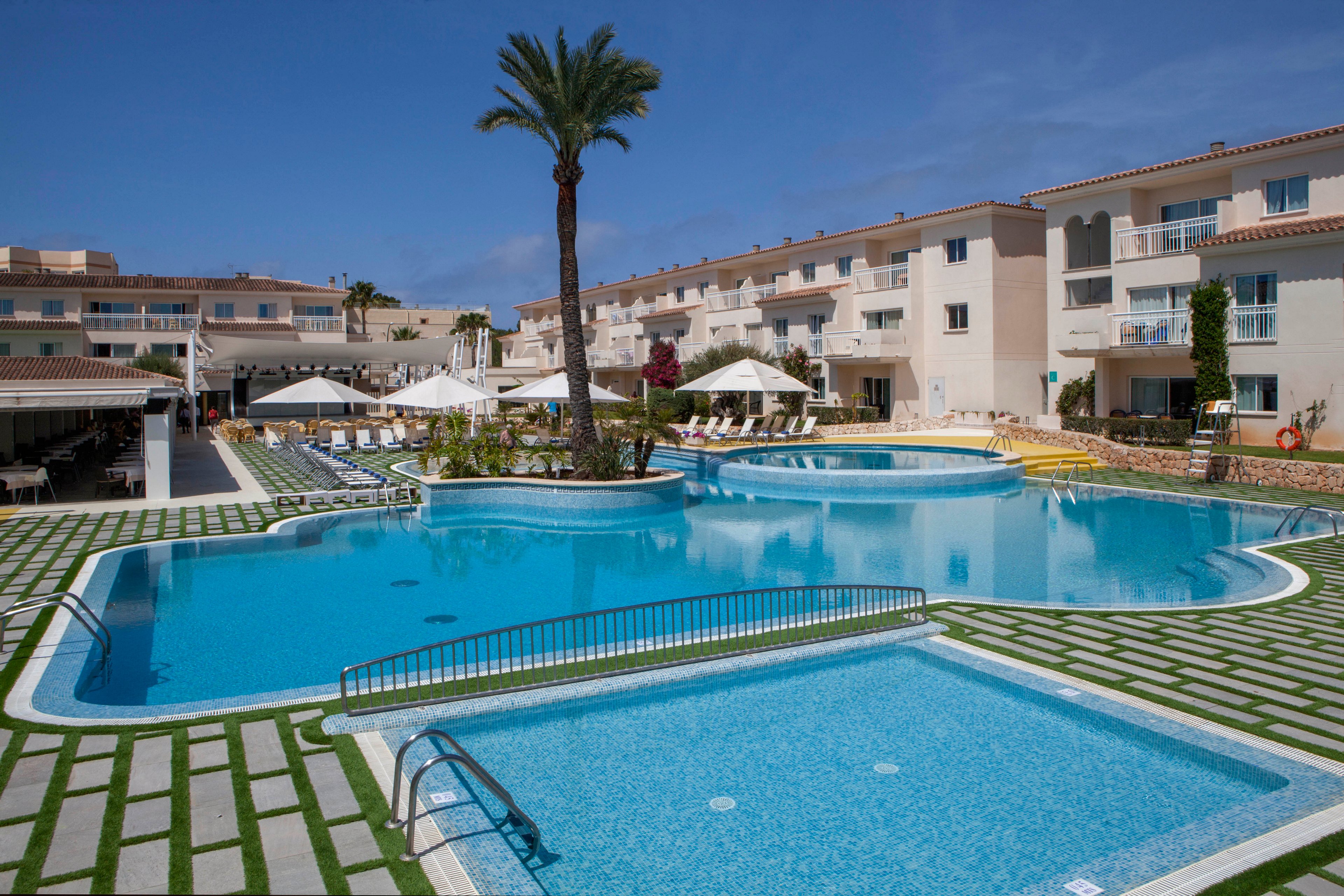 A large outdoor swimming pool is the central feature of this modern resort complex. White apartment buildings with balconies surround the pool, and sun loungers with umbrellas line the deck. The scene is bright and inviting, with clear blue skies and a single tall palm tree adding to the vacation atmosphere. No visible text or numbers are present in the image.