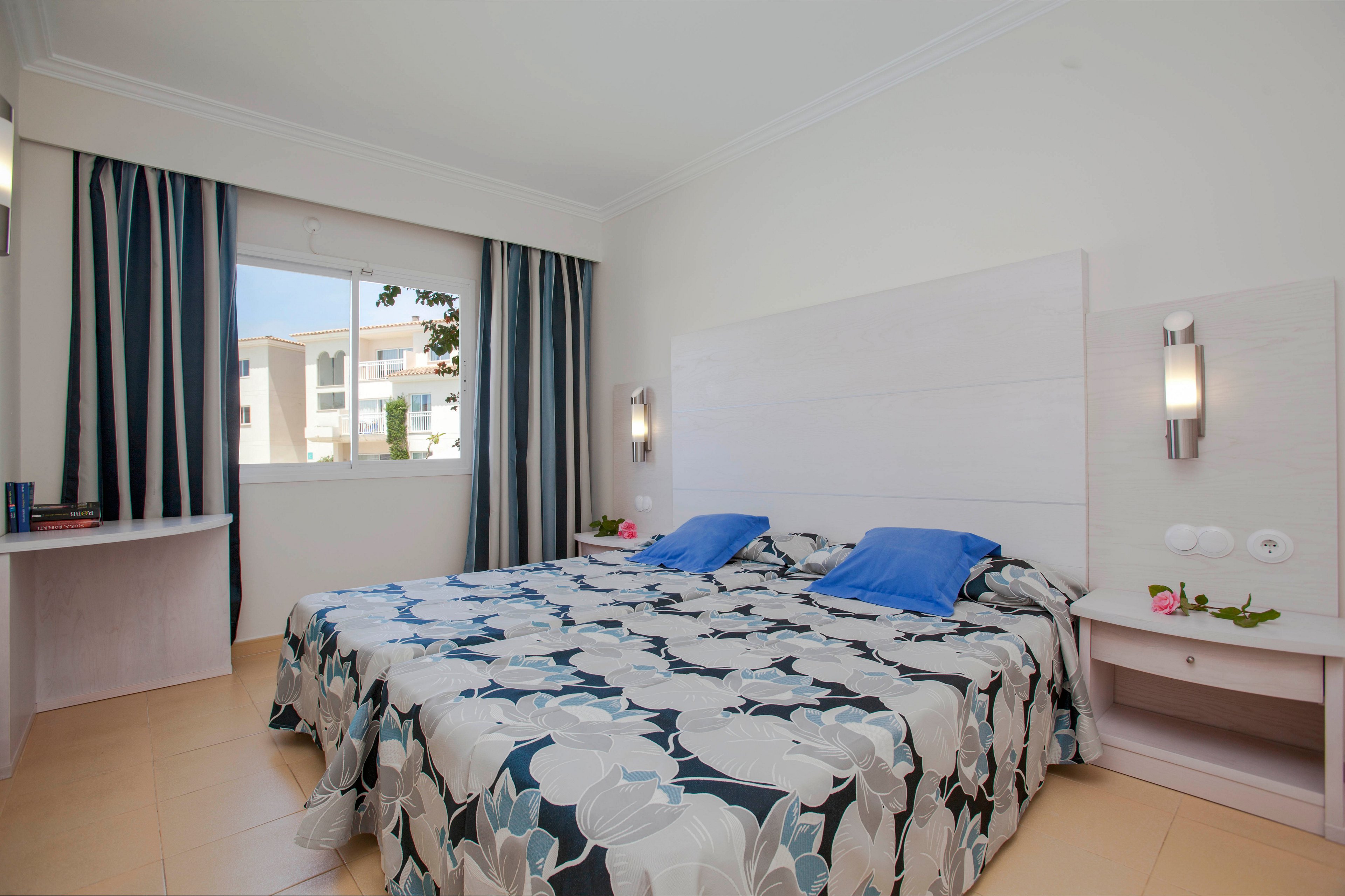 A modern hotel room features a double bed with a large floral-patterned bedspread and two blue pillows. The room is well-lit with natural light from a window, and includes simple white furniture and wall-mounted lights. The decor is minimal, with striped curtains and a clean, neutral palette. No visible text or numbers are present in the image.