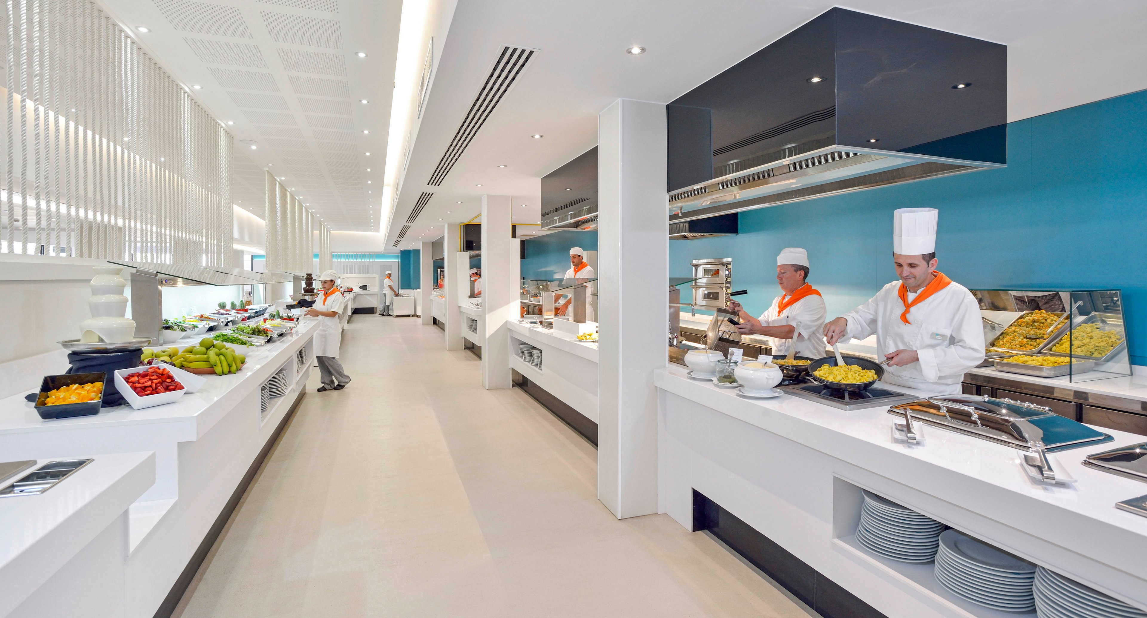 A spacious, contemporary buffet area features chefs in white uniforms and hats preparing and serving food. The setting is bright with clean white counters, blue accent walls, and a variety of fresh ingredients on display. The atmosphere is professional and inviting, with a focus on cleanliness and organization. Plates are neatly stacked, and the overall palette is white with pops of color from the food and decor.
