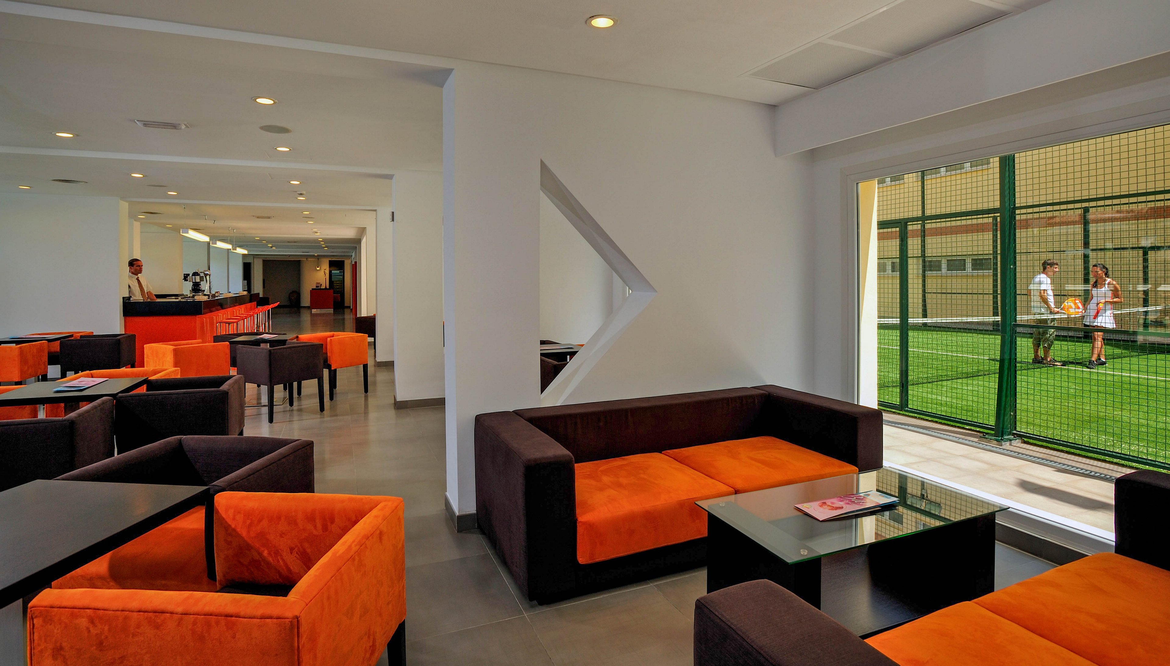 A contemporary lounge with orange and brown seating is shown adjacent to a glass wall, offering a clear view of an indoor sports court where people are playing. The setting is clean and well-lit, with geometric design elements and a relaxed atmosphere. The lounge features glass tables and a magazine, while the sports court adds an active, dynamic visual contrast. The image captures both leisure and activity in a modern environment.