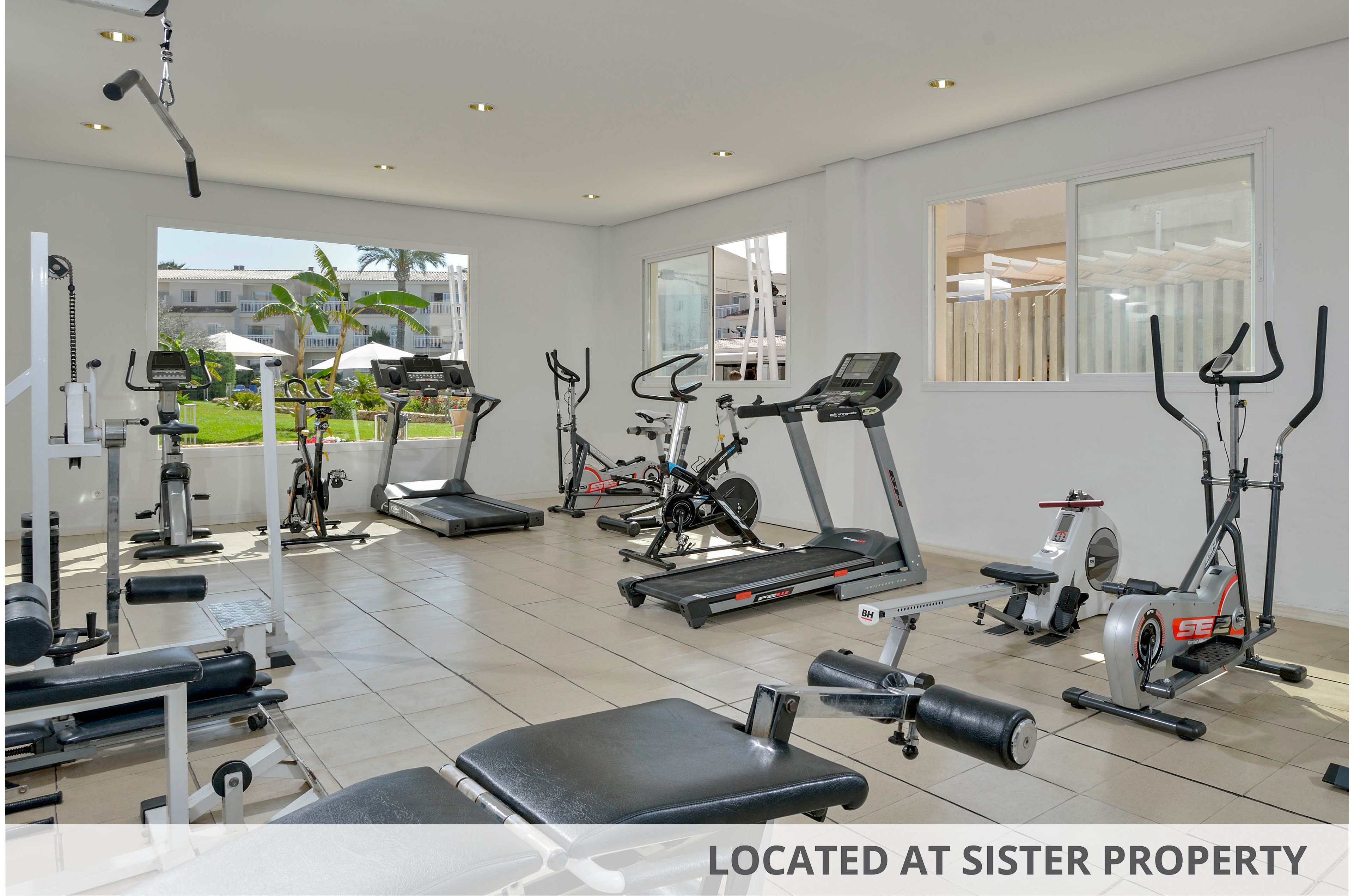 A bright, spacious gym features various exercise machines including treadmills, stationary bikes, and weight benches. The room is well-lit with large windows allowing natural light to fill the space. The clean, minimalist design creates an inviting atmosphere for fitness activities. No people are present, highlighting the equipment and open layout. The setting appears organized and ready for individual workouts.