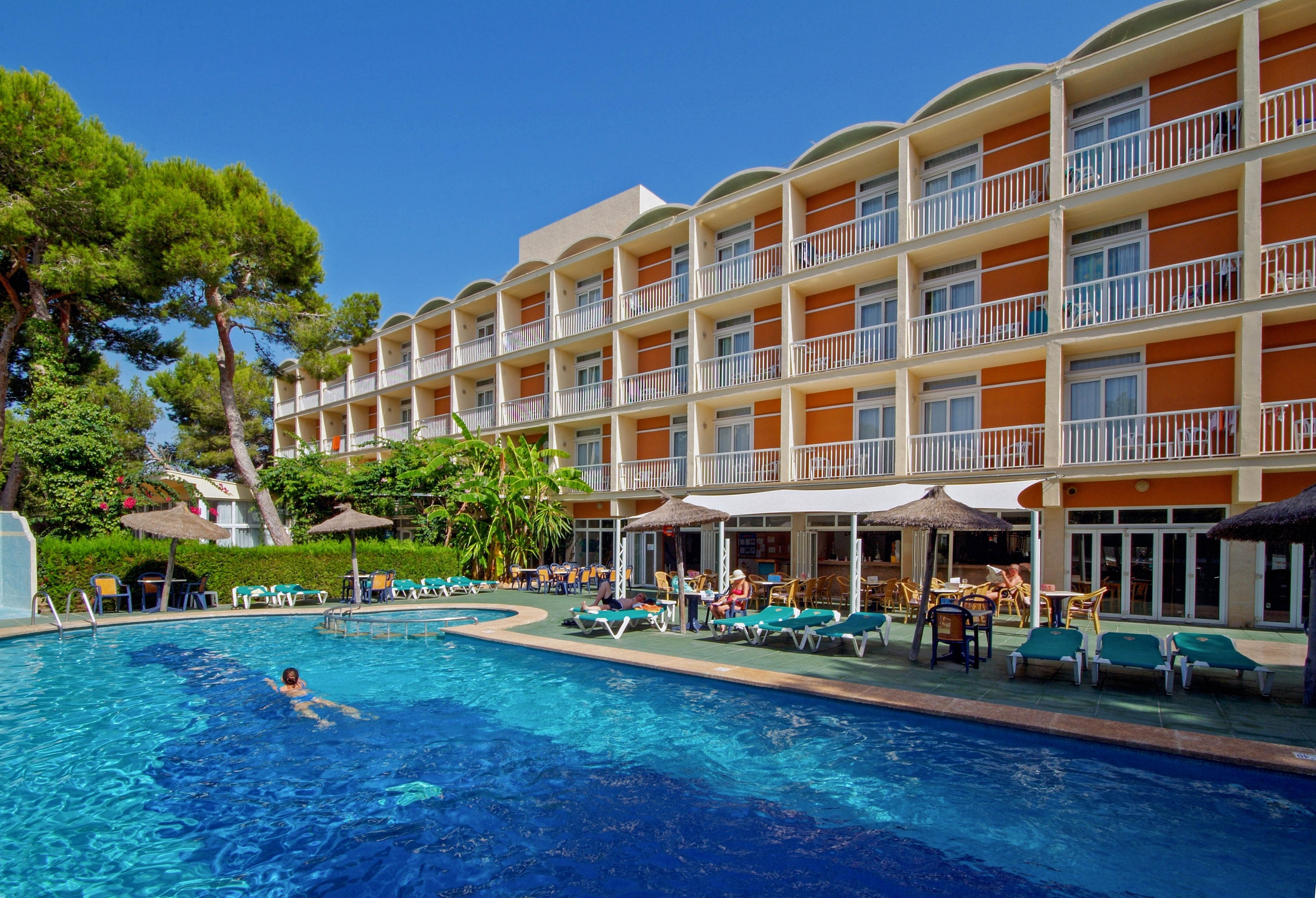 A multi-story hotel building with orange and white balconies overlooks a large outdoor swimming pool. Lounge chairs and sun umbrellas are arranged around the pool, with lush green trees in the background. One person is swimming in the clear blue water, while others relax poolside. The scene is bright and inviting, suggesting a warm, sunny day. No visible text or numbers are present in the image.