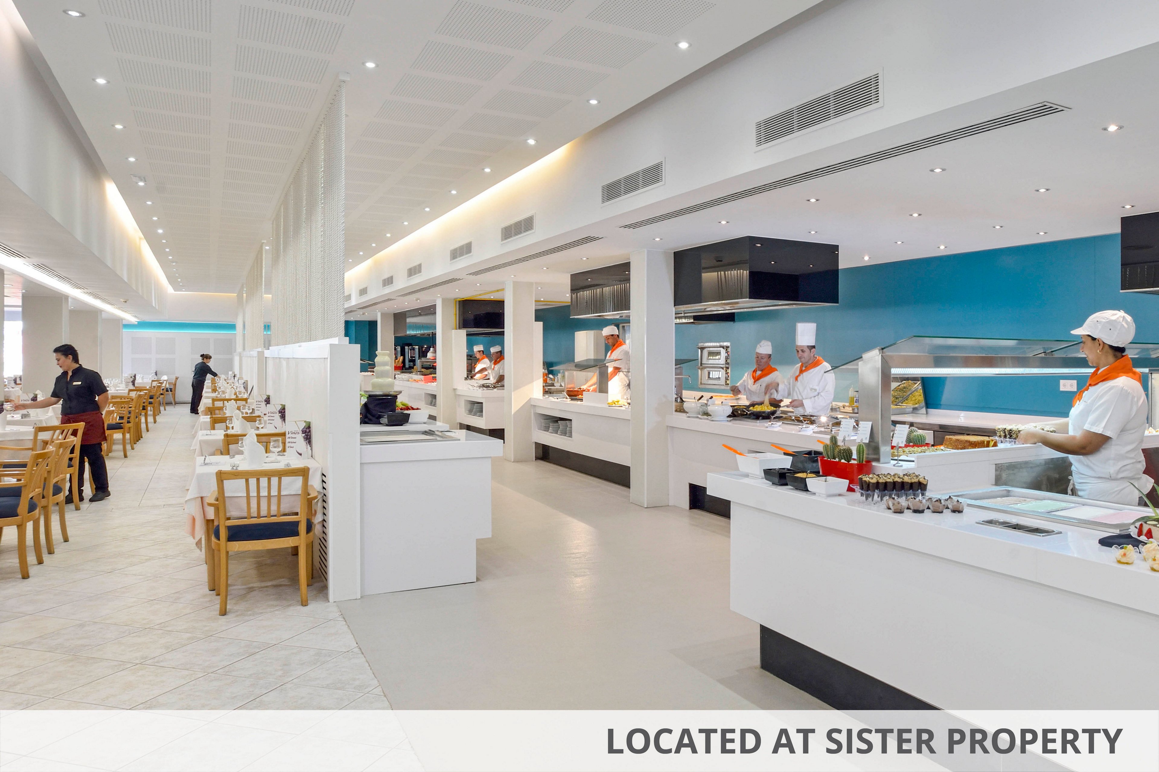 A spacious, brightly lit hotel buffet features chefs in white uniforms preparing and serving a variety of fresh dishes. The setting is clean and contemporary, with blue accent walls and neatly arranged tables in the dining area. The mood is inviting and professional, with a focus on fresh fruit and desserts displayed at the counter. Several staff members are visible behind the counters, while a few guests are seated in the background. No visible text or numeric values are present in the image.