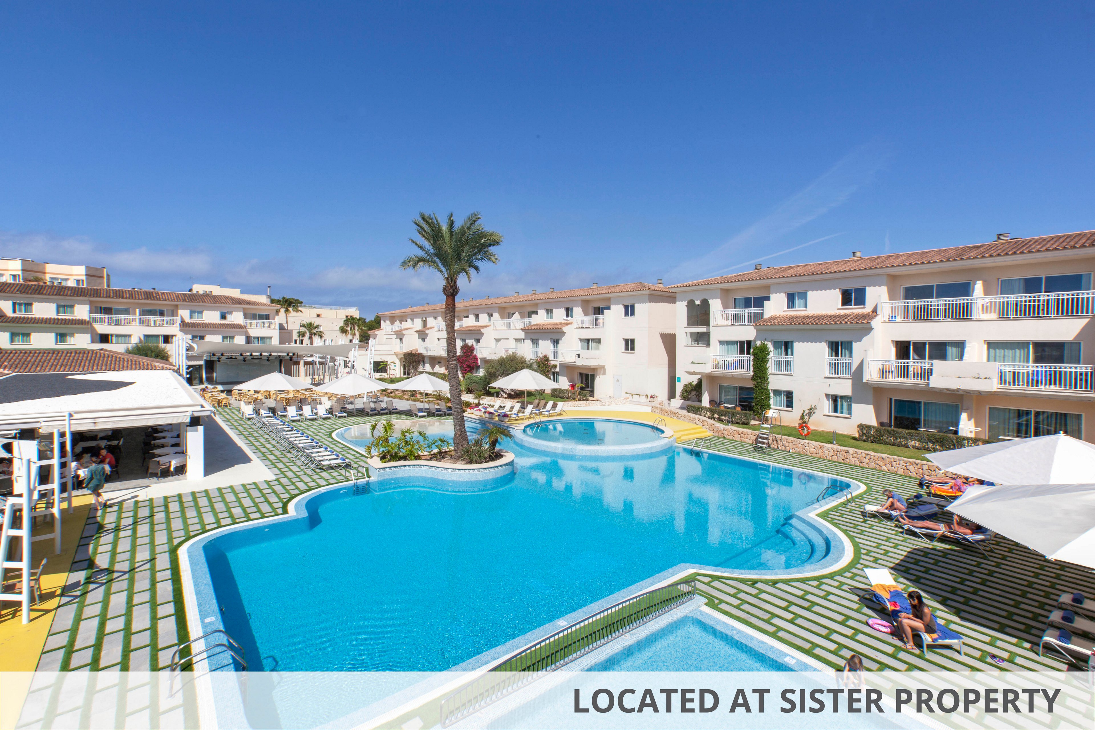 A large, uniquely shaped swimming pool is the central feature of this sunny resort complex. Modern white apartment buildings with balconies surround the pool, while lounge chairs and umbrellas provide shaded relaxation spots. A tall palm tree stands out near the pool, and a clear blue sky enhances the bright, inviting atmosphere. Several people are seen enjoying the outdoor amenities, contributing to a lively vacation mood.