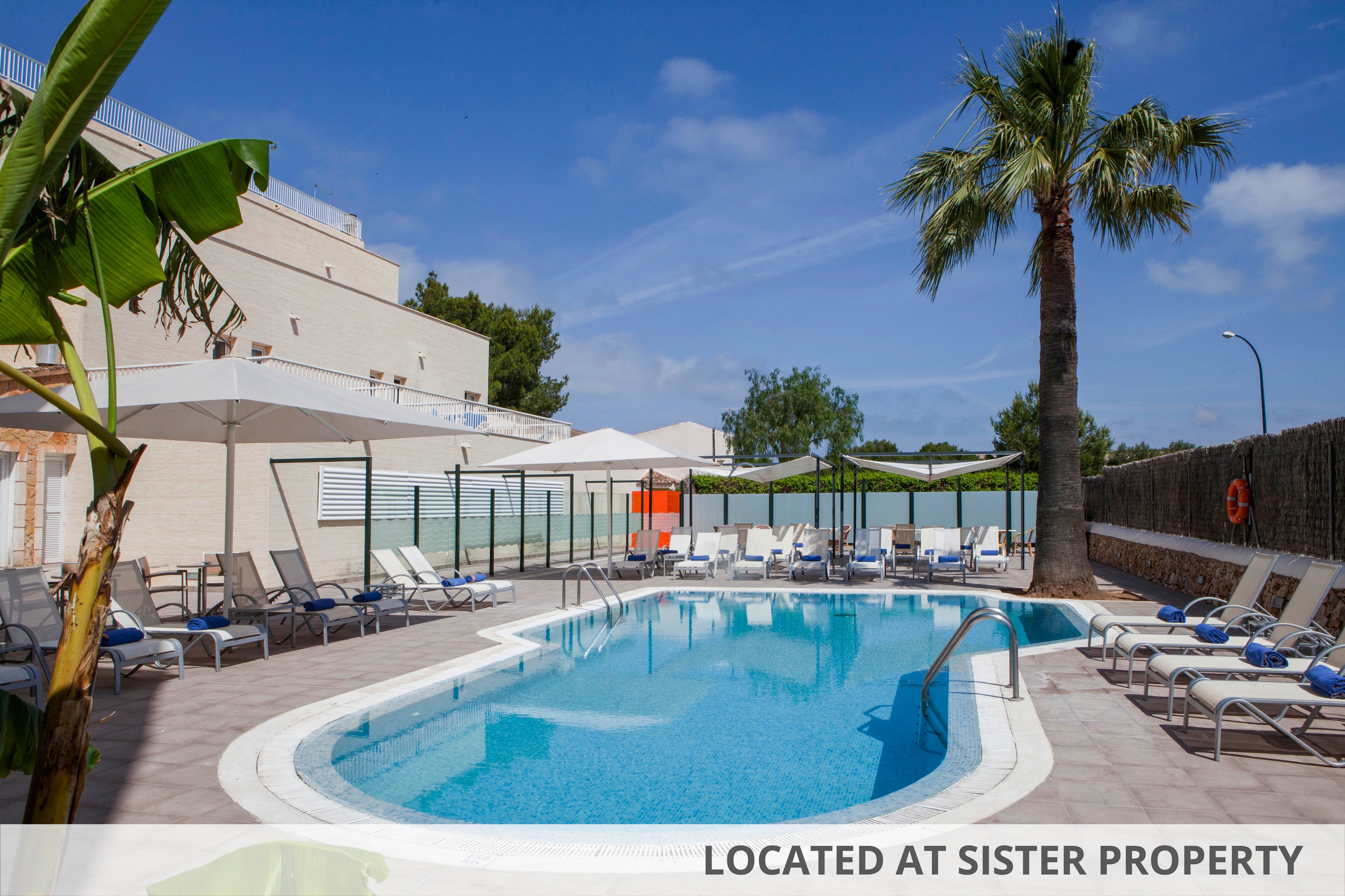 A clear blue swimming pool is the central focus of this outdoor hotel area, surrounded by numerous lounge chairs and shaded umbrellas. The setting is bright and inviting, with a tall palm tree and tropical plants adding to the relaxing atmosphere. The sky is mostly clear, and the pool deck is paved with light tiles, creating a clean and modern look. No people are present, emphasizing tranquility and leisure.