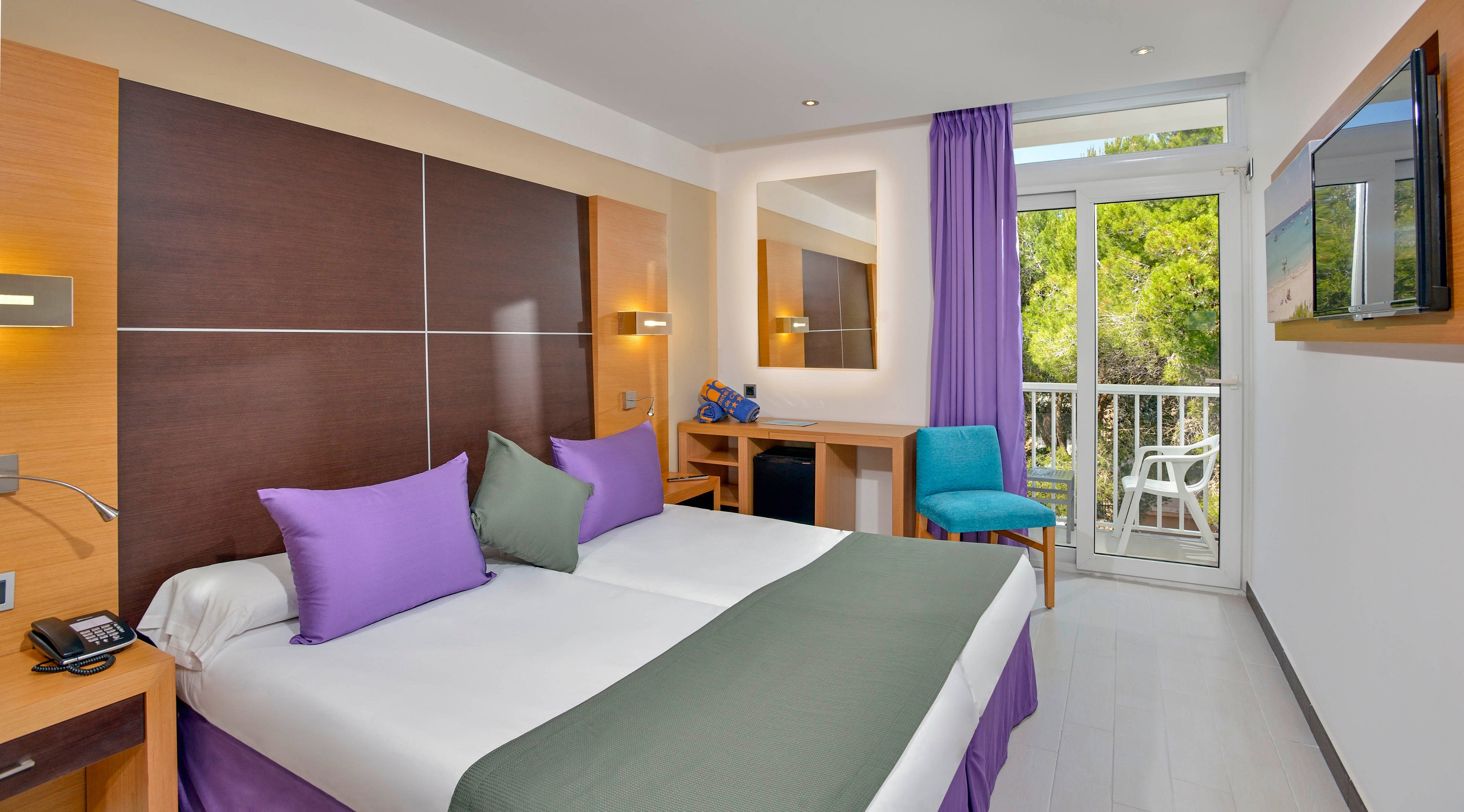 A contemporary hotel room features two twin beds pushed together, adorned with purple and gray bedding and pillows. The space includes a wooden desk, a turquoise chair, and a wall-mounted television. Large windows and a glass door open to a balcony with a view of green trees, allowing natural light to fill the room. The overall mood is bright and inviting, with a clean and organized appearance.