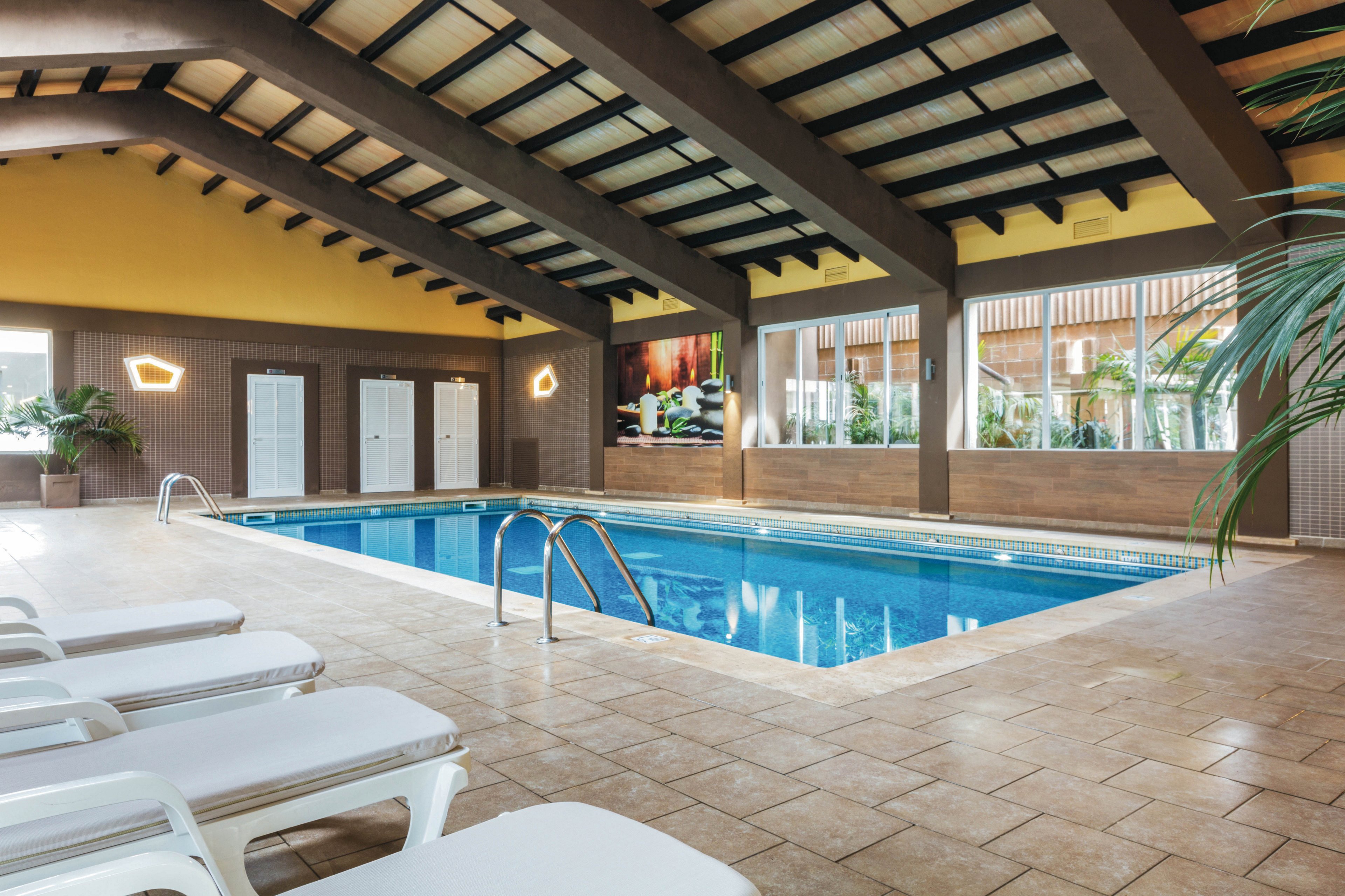 A spacious indoor swimming pool is featured in a modern recreational facility. The pool area includes several white lounge chairs, tiled flooring, and large windows that allow natural light to fill the space. The ceiling is constructed with exposed beams, and the walls are decorated with geometric lights and tropical plants. No people are present, creating a calm and inviting atmosphere.