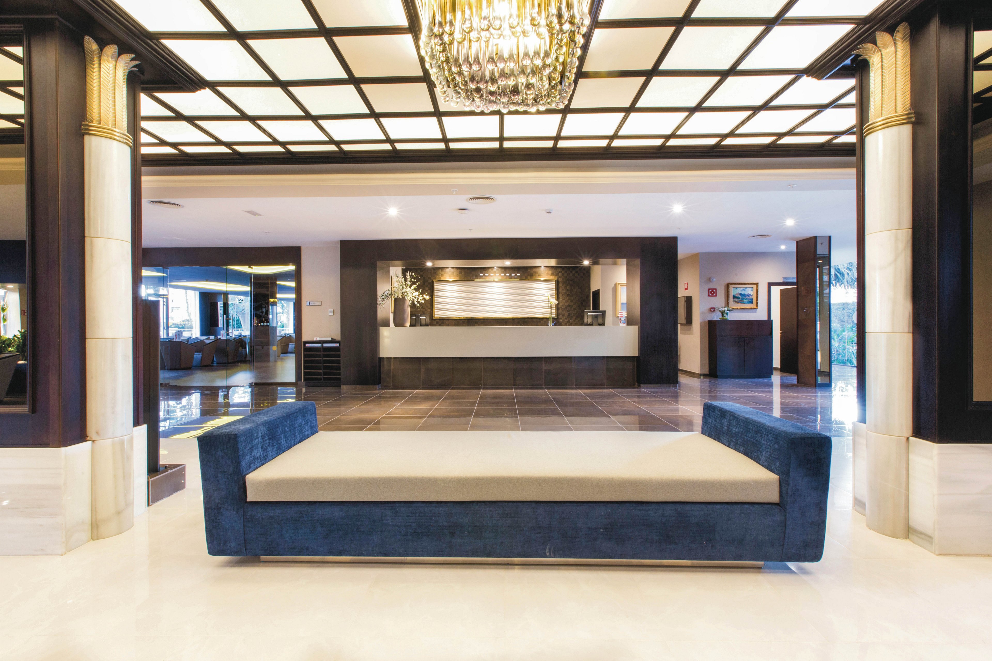 A spacious hotel lobby features a contemporary blue velvet bench in the foreground and a well-lit reception desk in the background. The setting is upscale, with a geometric ceiling light fixture and polished marble floors enhancing the luxurious atmosphere. The color palette is neutral with accents of gold and navy, creating a welcoming and sophisticated mood. No visible people or text are present in the image.