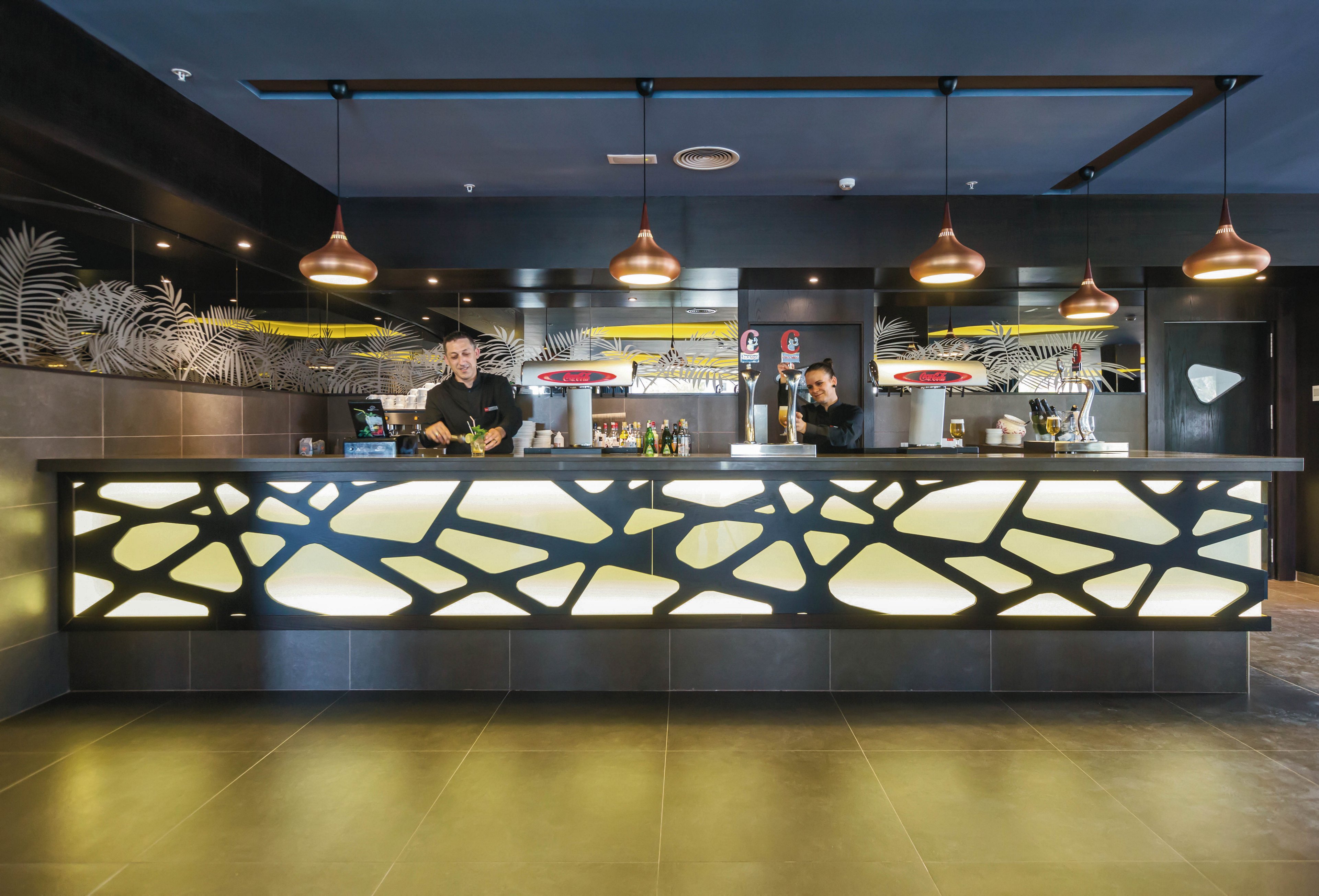 A contemporary hotel reception area features a striking illuminated front desk with geometric patterns. Two staff members stand behind the counter, ready to assist guests. The setting is stylish and modern, with pendant lighting and decorative wall accents. The overall mood is professional and welcoming, with a neutral and warm color palette.