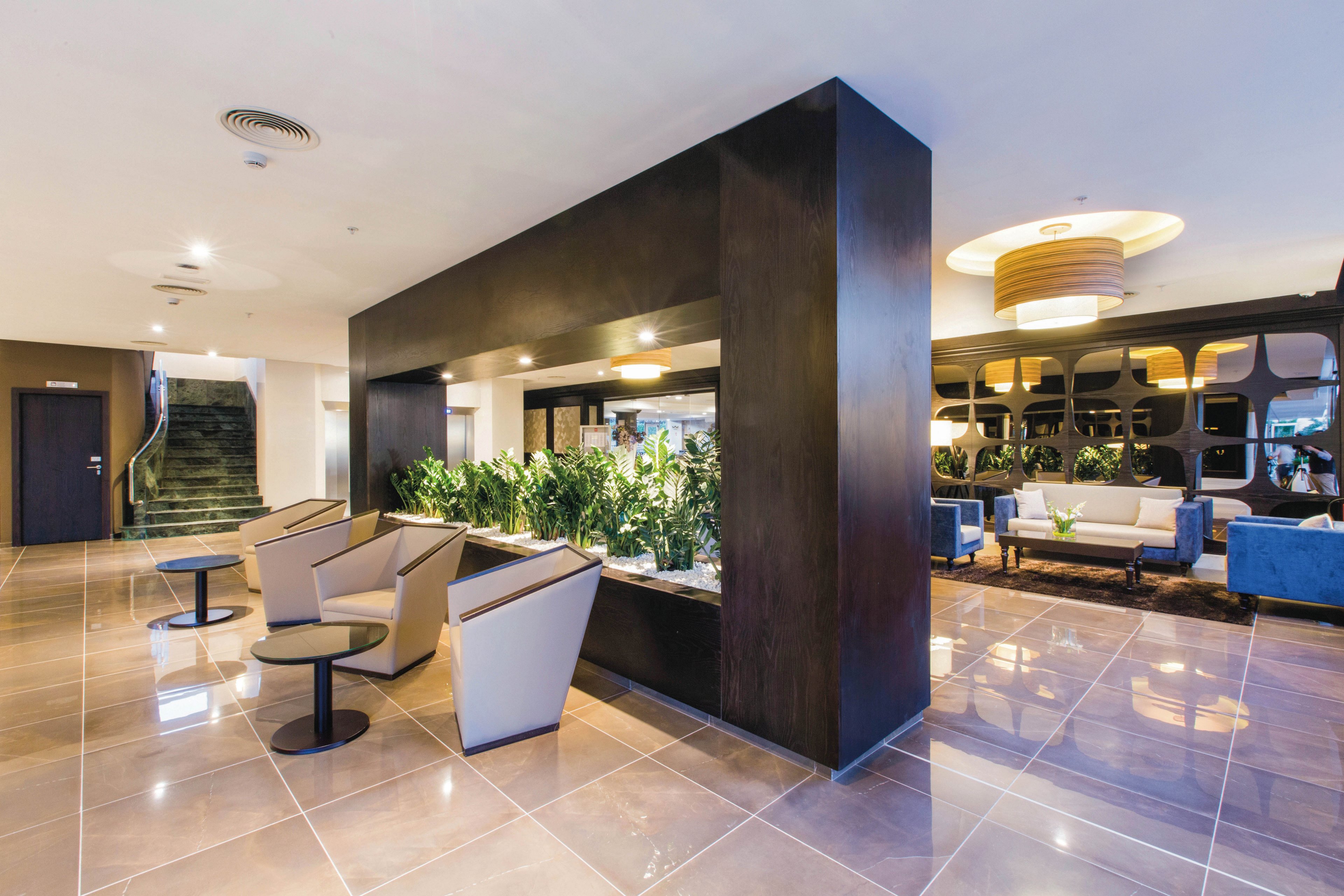 A spacious hotel lobby features modern armchairs and tables arranged around a central divider with lush green plants. The setting is bright and welcoming, with polished tile floors and stylish lighting fixtures. Blue sofas and geometric wall accents add a touch of sophistication to the lounge area. No visible people or text are present in the image.