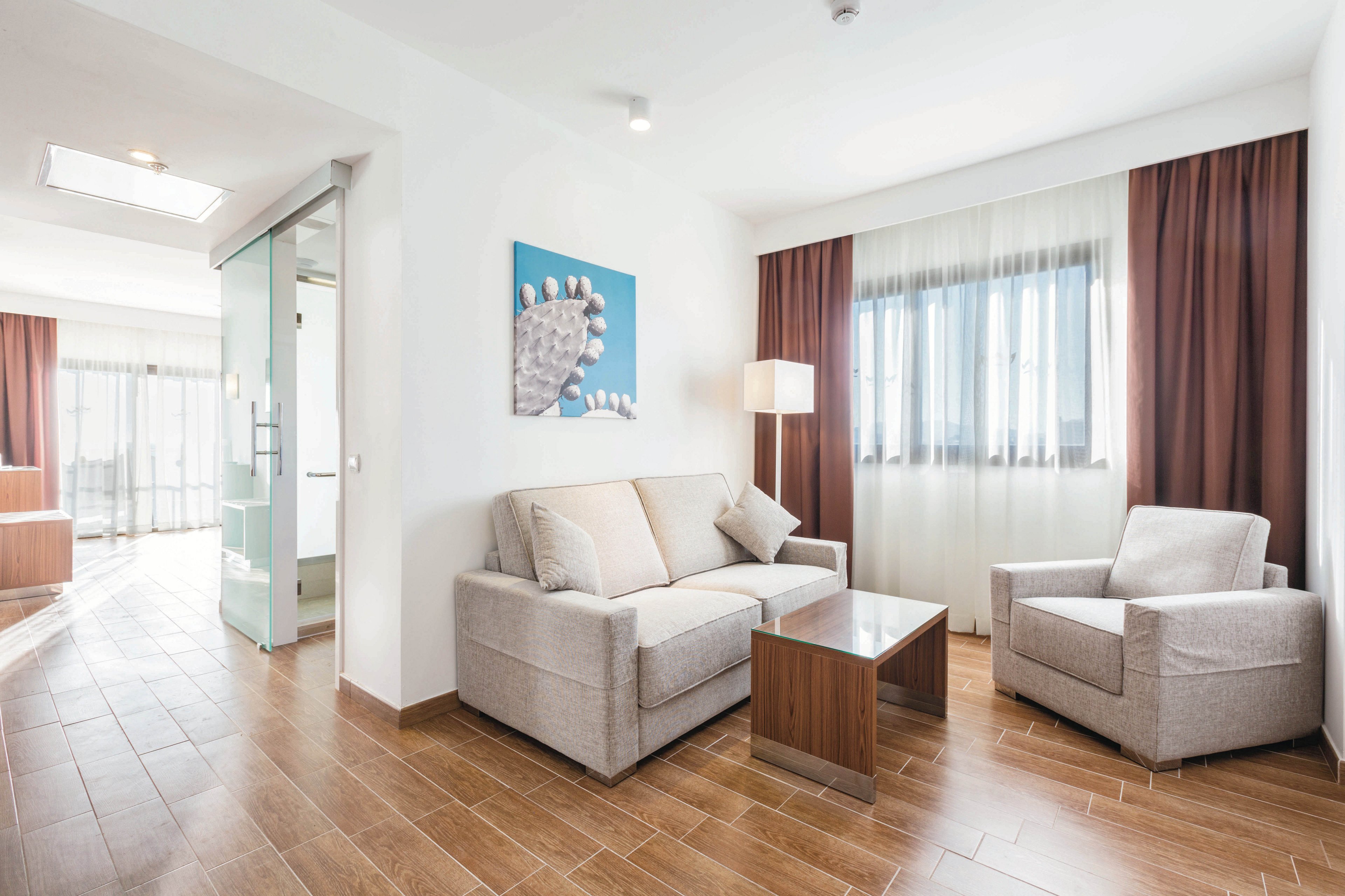 A bright, modern hotel suite features a neutral-toned sofa and armchair arranged around a glass coffee table. The space is illuminated by natural light from a large window with sheer curtains, complemented by a floor lamp. Wooden flooring and minimalist artwork add warmth and style to the clean, contemporary setting. The suite includes a visible bathroom area separated by a glass partition.