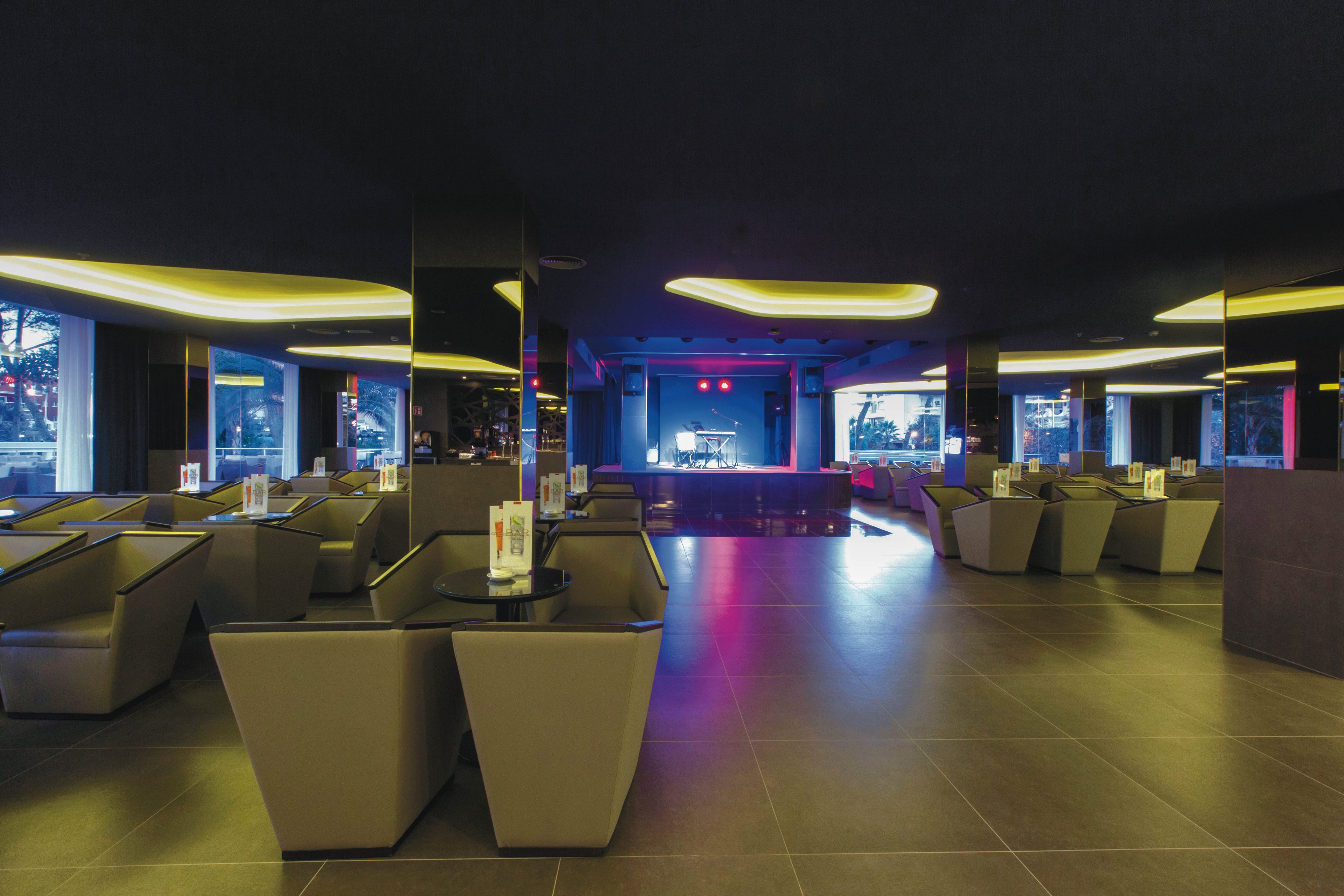 A spacious, contemporary lounge features geometric seating arrangements and a central stage area. The setting is illuminated by soft, recessed ceiling lights and colored accent lighting near the stage. Large windows allow natural light to filter in, while the overall palette is neutral with dark flooring and muted furniture. The atmosphere suggests a relaxed, upscale environment suitable for social gatherings or live performances. No visible text or numbers are present in the image.