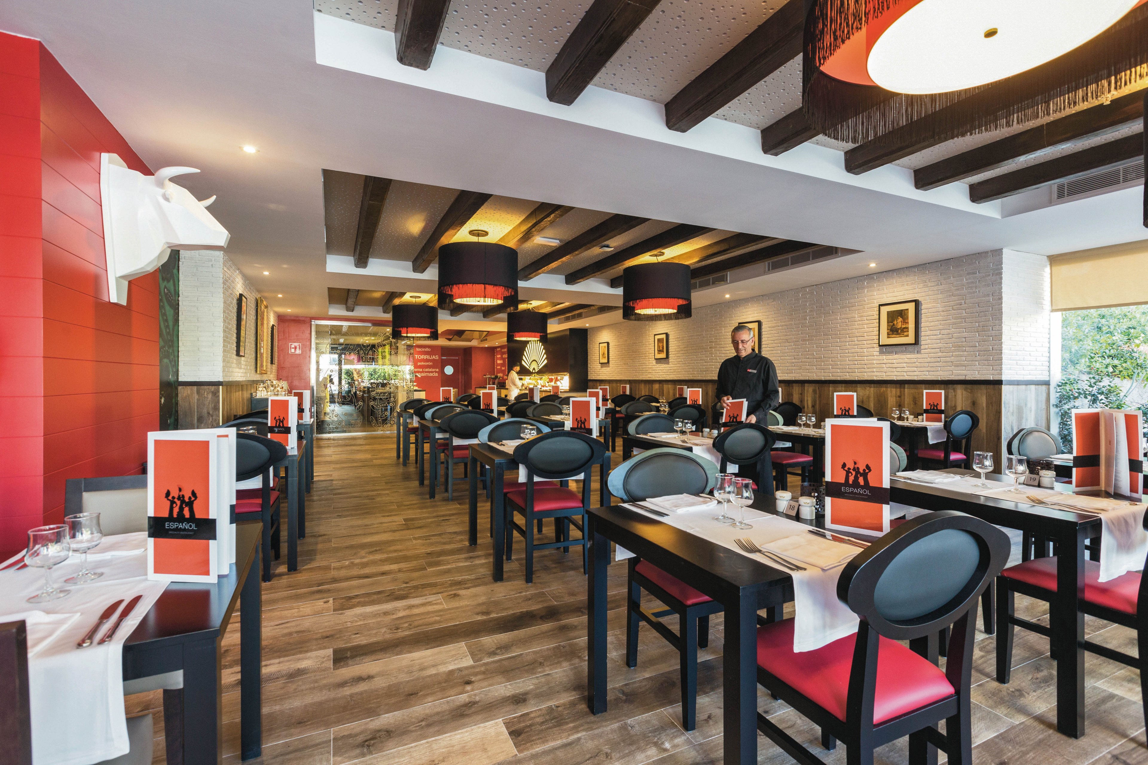 A contemporary restaurant dining area features neatly arranged tables with red and black chairs. A single male staff member stands near the center, ready to serve guests. The setting is well-lit with large pendant lights, wooden beams, and decorative wall art. Table settings include menus with silhouetted figures and white napkins. The overall mood is inviting and stylish, with a warm color palette.