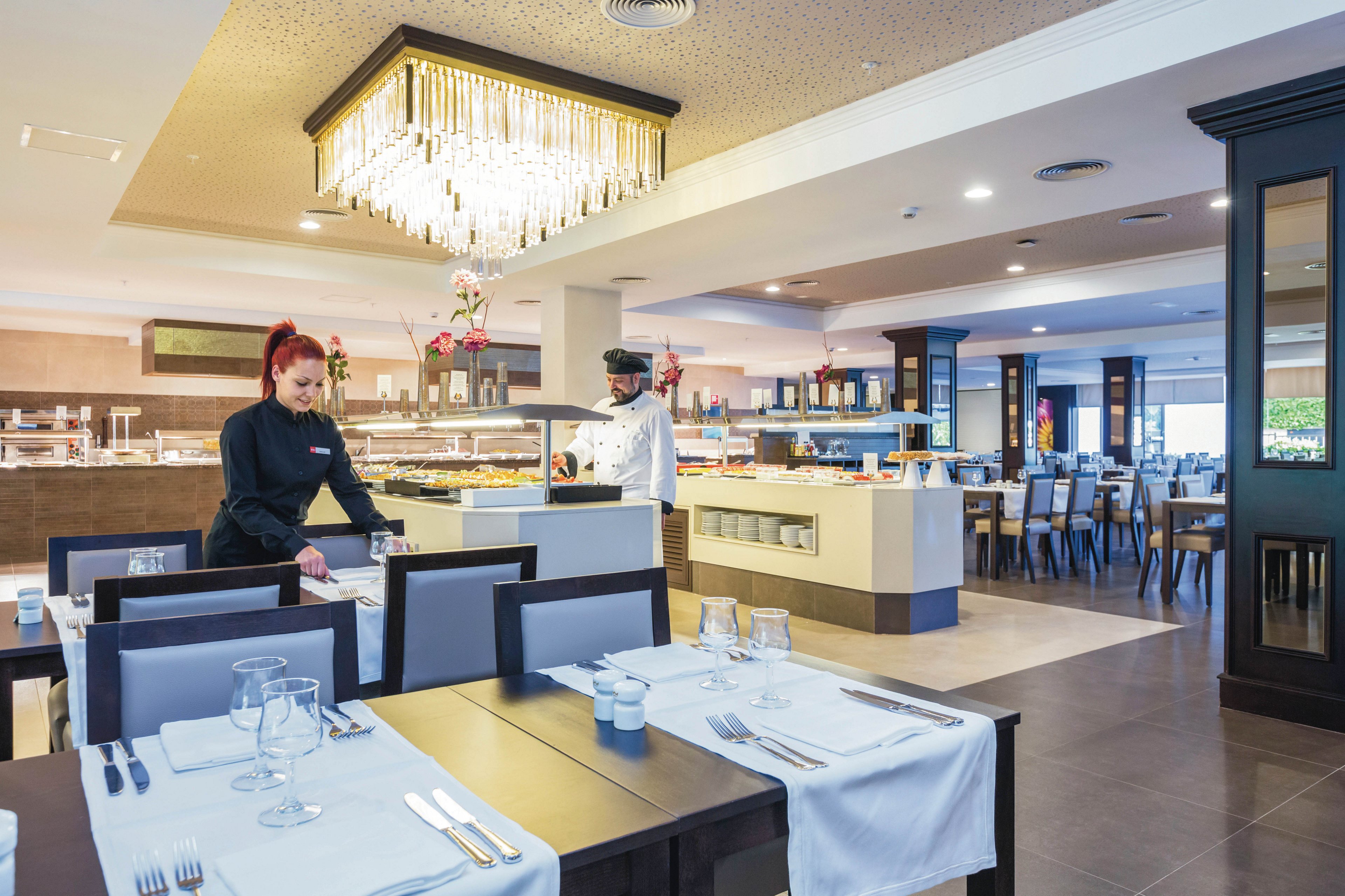 A spacious, contemporary restaurant dining area is shown with neatly set tables and elegant decor. Two staff members, one in a black uniform and another in a white chef's outfit, are preparing the space for service. The setting features a large chandelier, neutral color palette, and a buffet area in the background. The atmosphere is clean, organized, and welcoming, with no visible text or numbers present.