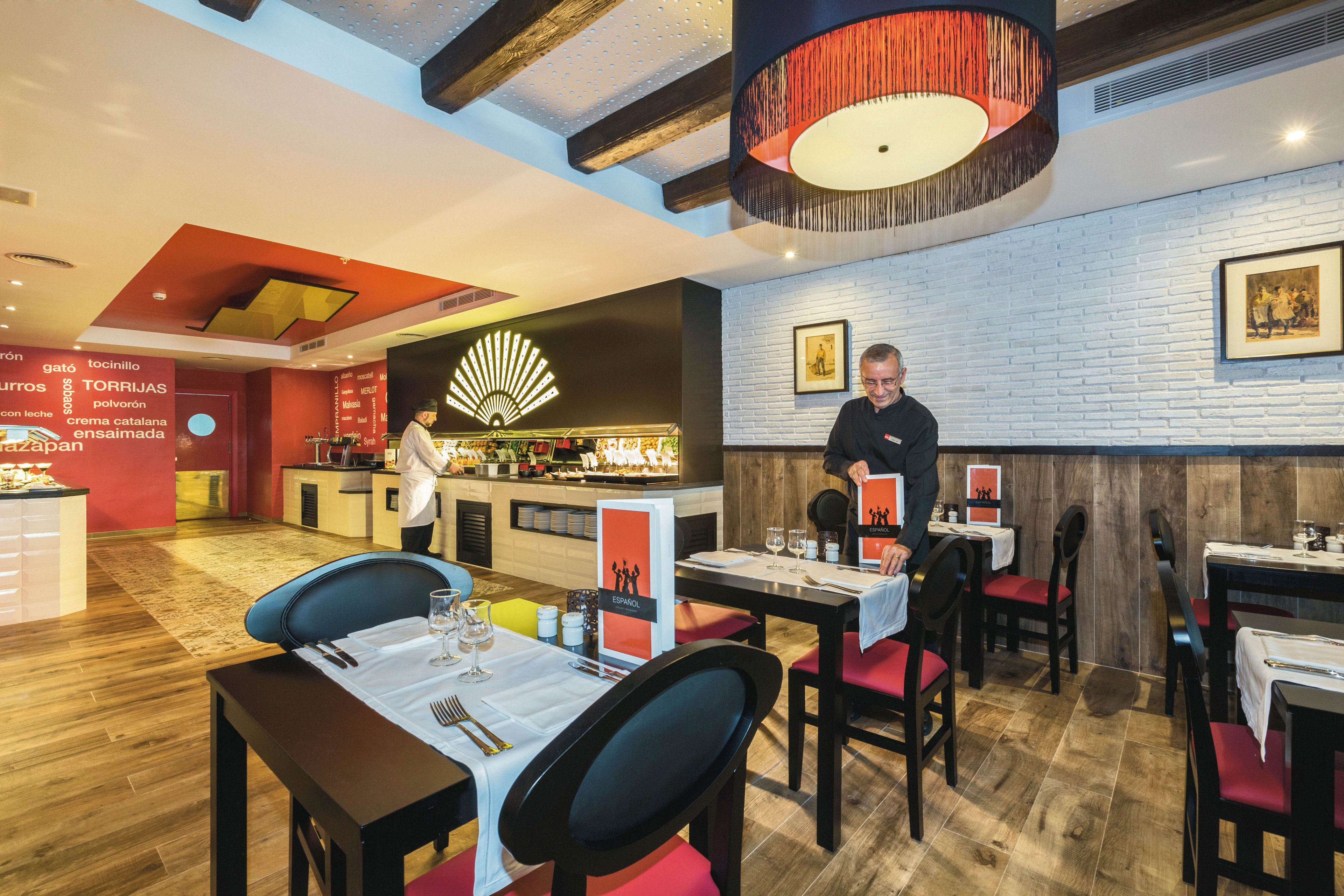 A contemporary restaurant dining area features wooden floors, red and black chairs, and decorative wall art. A male staff member is setting tables, while another person is seen near the buffet area. The ambiance is warm with soft lighting and a stylish ceiling lamp, and visible menus are placed on the tables. The setting suggests a casual yet refined dining experience.