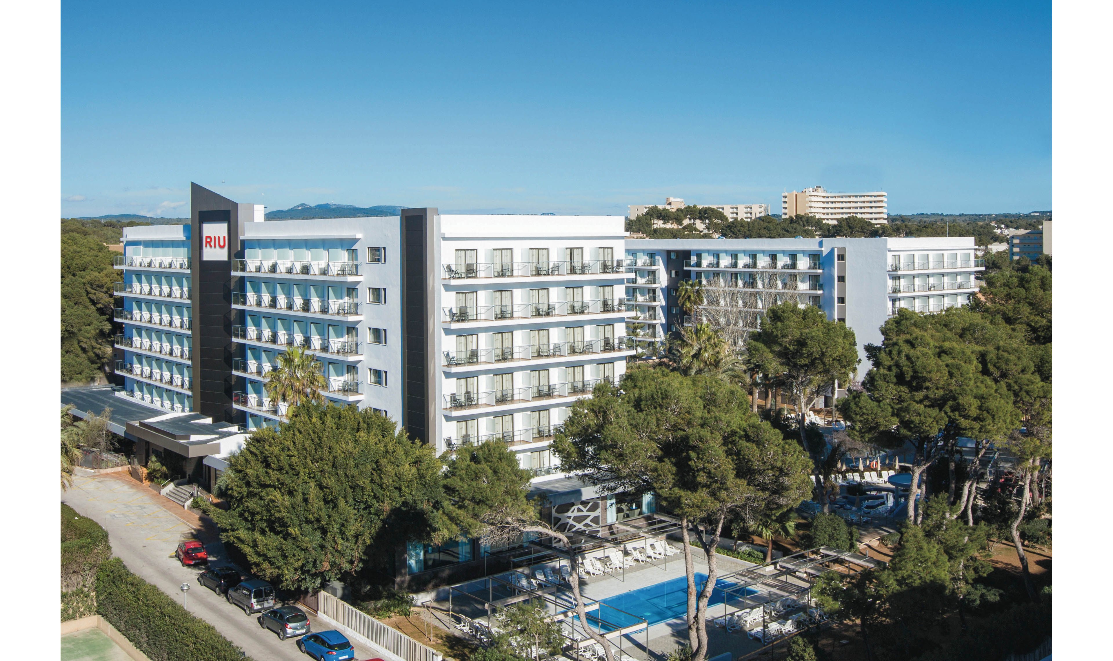 A large, contemporary RIU hotel building is shown surrounded by lush trees and greenery. The setting features a prominent outdoor swimming pool with sun loungers and umbrellas, indicating a leisure or vacation environment. The hotel has multiple floors with balconies, and the RIU name is clearly visible on the building facade. The sky is clear, suggesting a bright and inviting atmosphere.