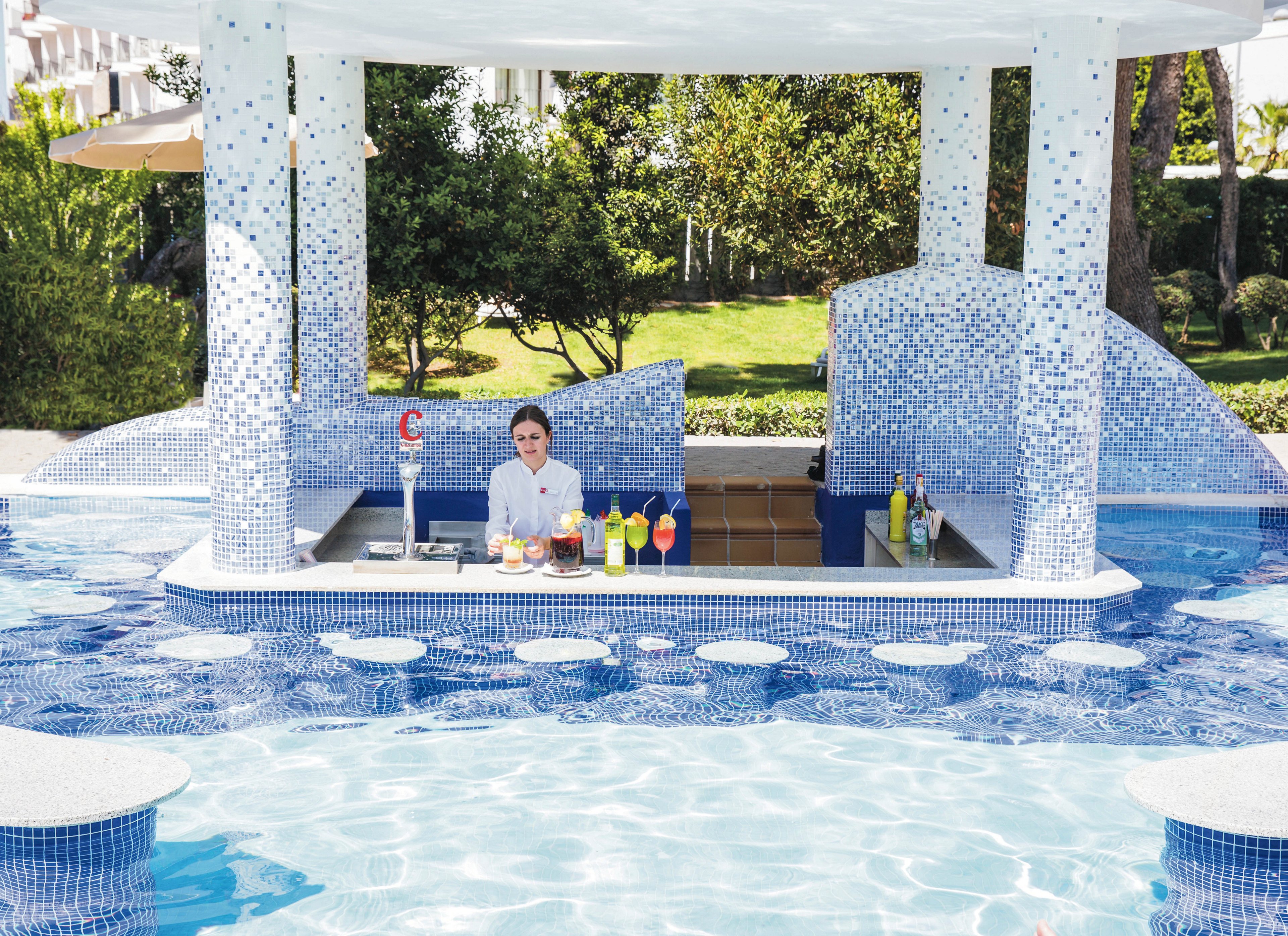 A swim-up bar is set in a blue-tiled pool, featuring a bartender in a white uniform preparing and serving a variety of colorful beverages. The bar is shaded by a white canopy and surrounded by lush greenery, creating a relaxing resort atmosphere. Several vibrant cocktails and bottles are displayed on the counter, enhancing the inviting vacation vibe. The scene is bright and summery, with a clean and modern design.