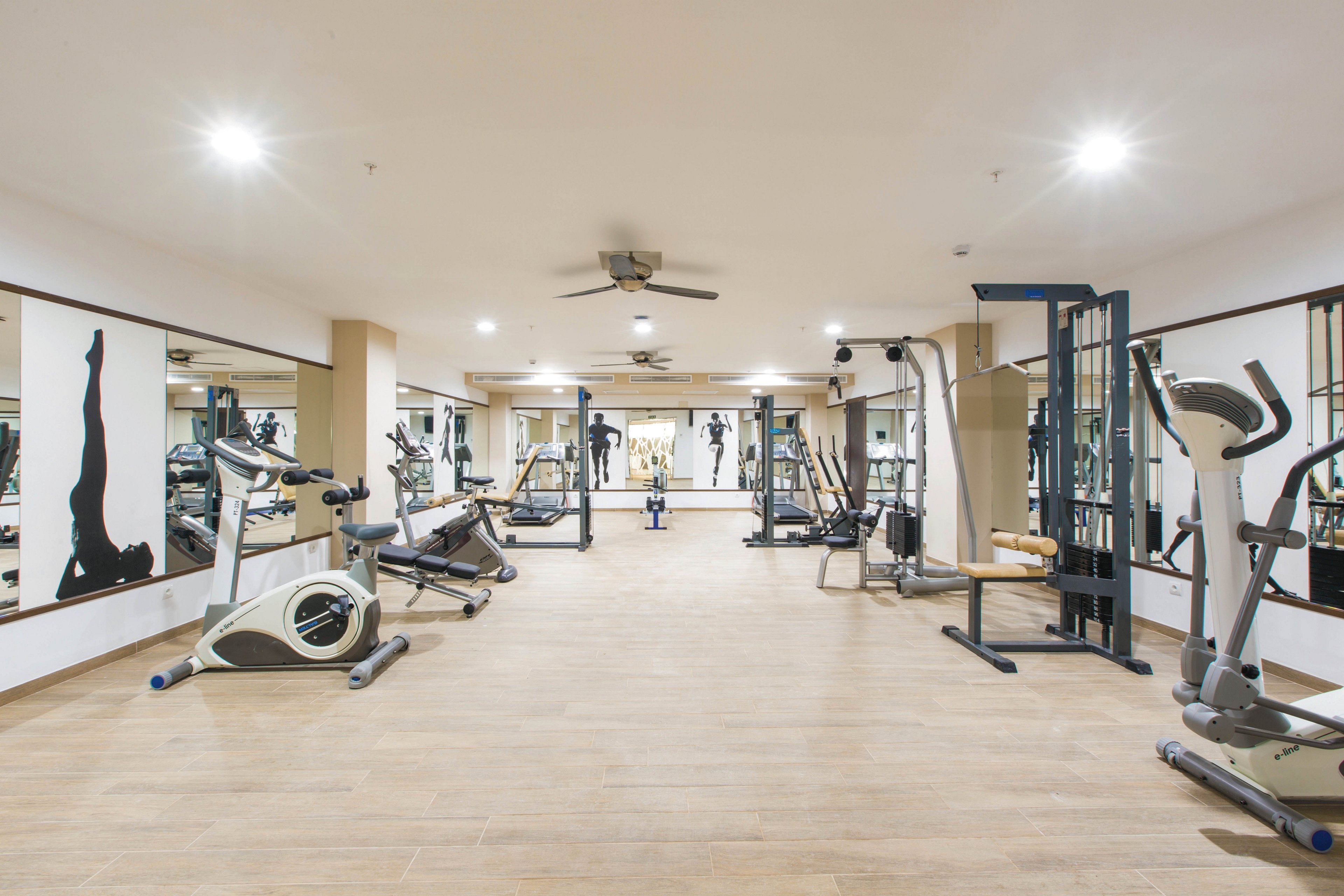 A spacious gym features various exercise machines including treadmills, stationary bikes, and weight equipment. The room is well-lit with ceiling lights and has large mirrors along the walls, enhancing the sense of space. The wooden flooring and neutral color palette create a clean, contemporary atmosphere. Silhouettes of athletes are visible on the wall mirrors, adding a dynamic visual element. No people are present in the scene.