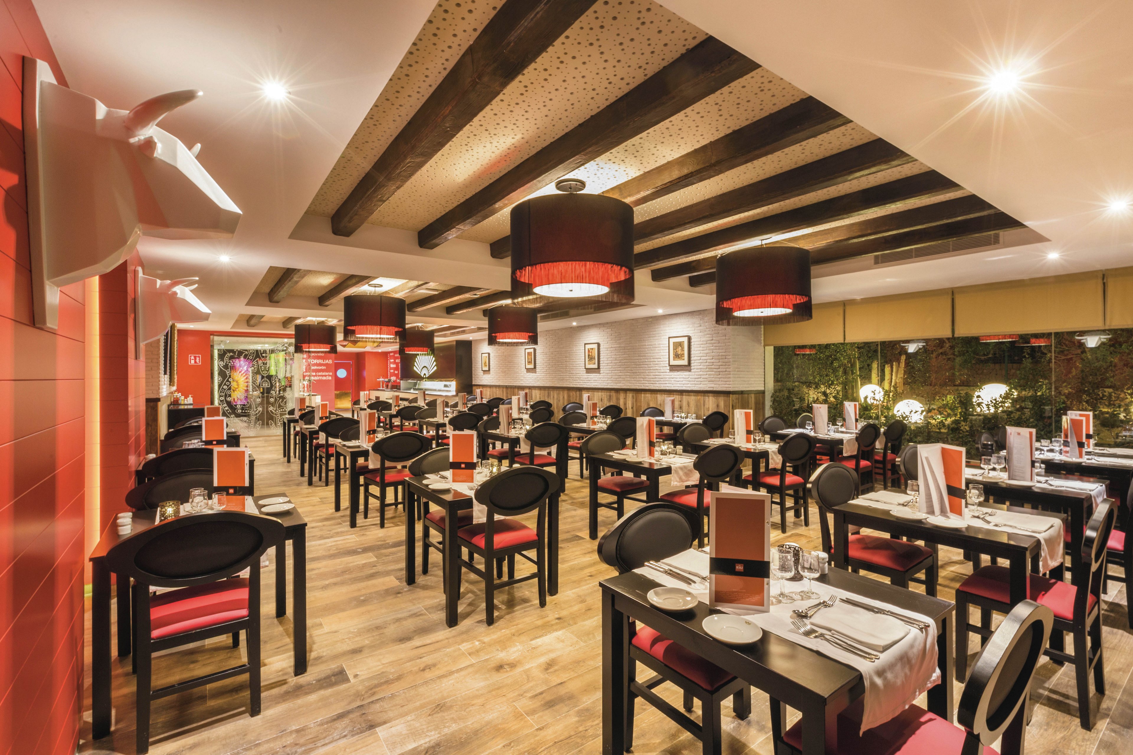 A contemporary restaurant dining area features neatly arranged tables and chairs with red and black accents. The setting is well-lit with stylish pendant lights and wooden ceiling beams, creating a warm and inviting atmosphere. Table settings include menus, plates, and glasses, while framed artwork decorates the walls. No visible people are present, emphasizing the clean and organized environment.