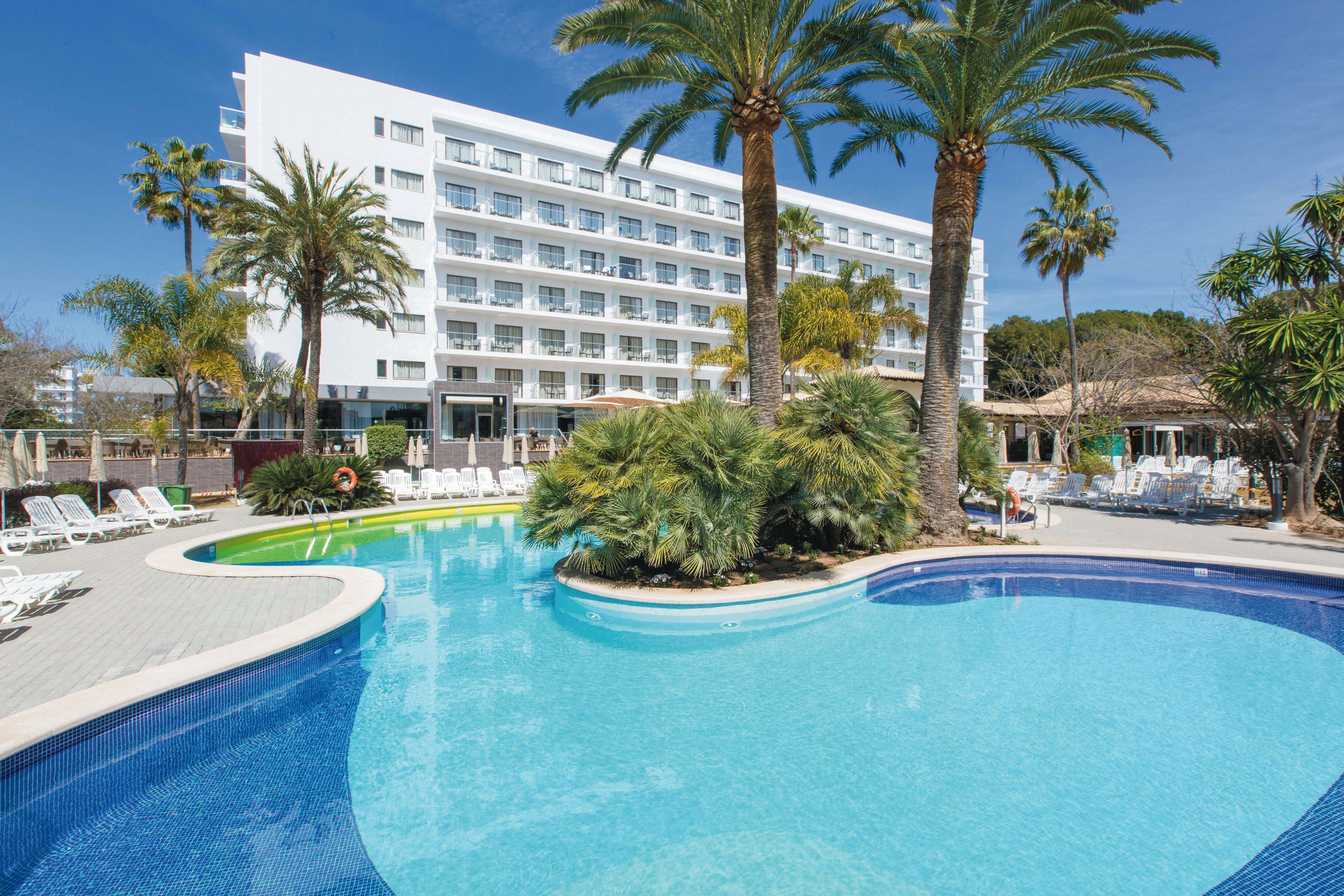 A contemporary multi-story hotel stands behind a large outdoor swimming pool surrounded by lush palm trees. The pool area features clear blue water, sun loungers, and a relaxed resort atmosphere. The sky is clear, enhancing the bright and inviting mood of the scene. No visible text or numbers are present in the image.