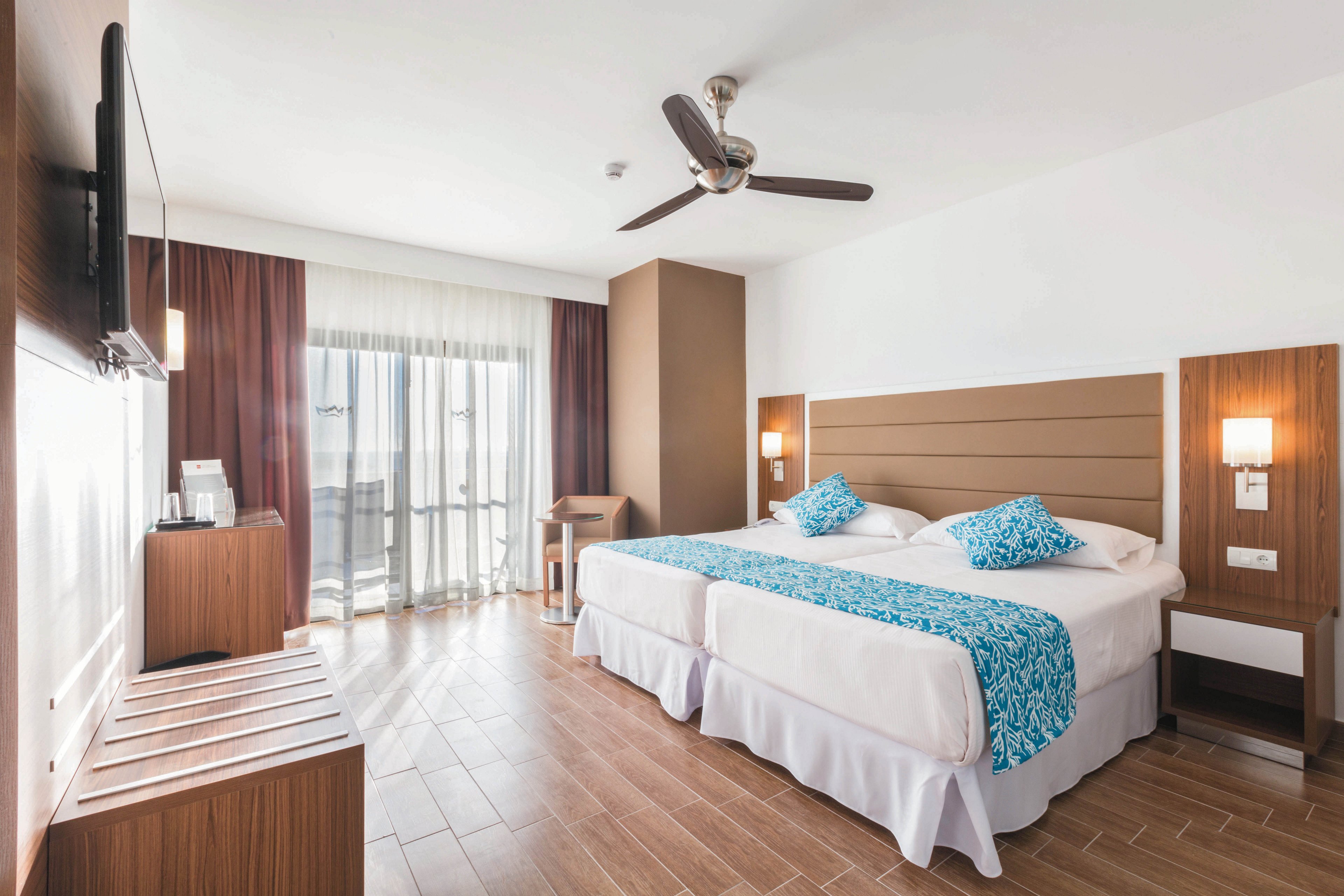 A bright, contemporary hotel room features two twin beds with blue patterned runners and pillows. The space includes wood accents, a ceiling fan, wall-mounted TV, and a large window with sheer curtains leading to a balcony. The room is well-lit with natural light, and the decor is minimalist with a neutral and blue color palette. No visible people or explicit text are present in the image.