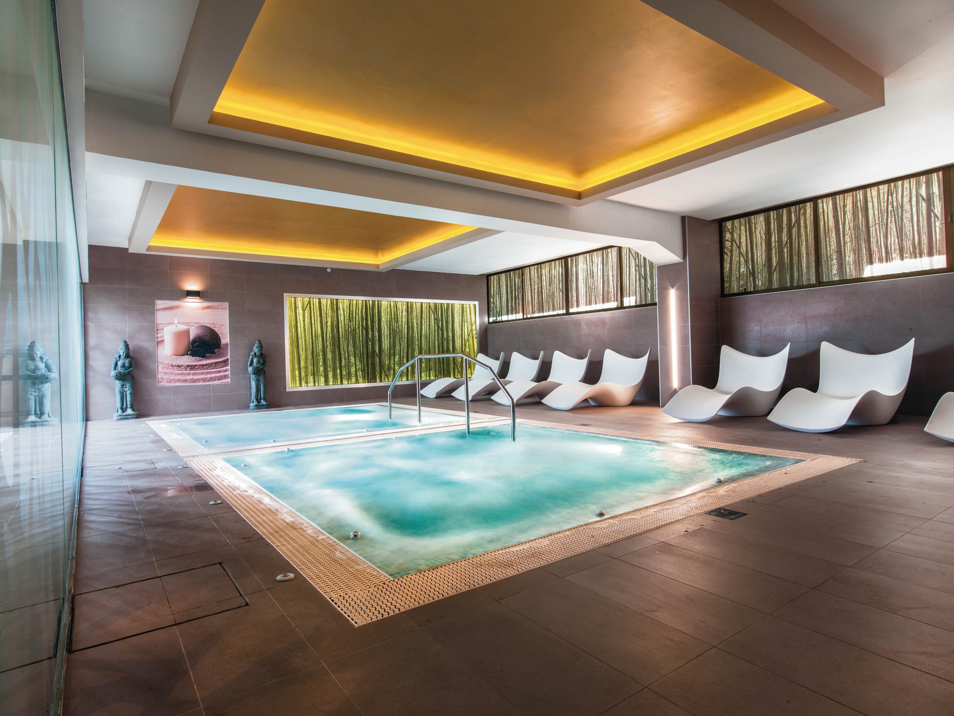 A contemporary indoor spa pool is the main focus, surrounded by sleek white lounge chairs and decorative statues. The setting features warm recessed lighting, large windows with bamboo visuals, and a tranquil atmosphere. The pool area is clean and inviting, with a minimalist design and a relaxing ambiance. A poster with a visible circular graphic and some text is displayed on the wall.