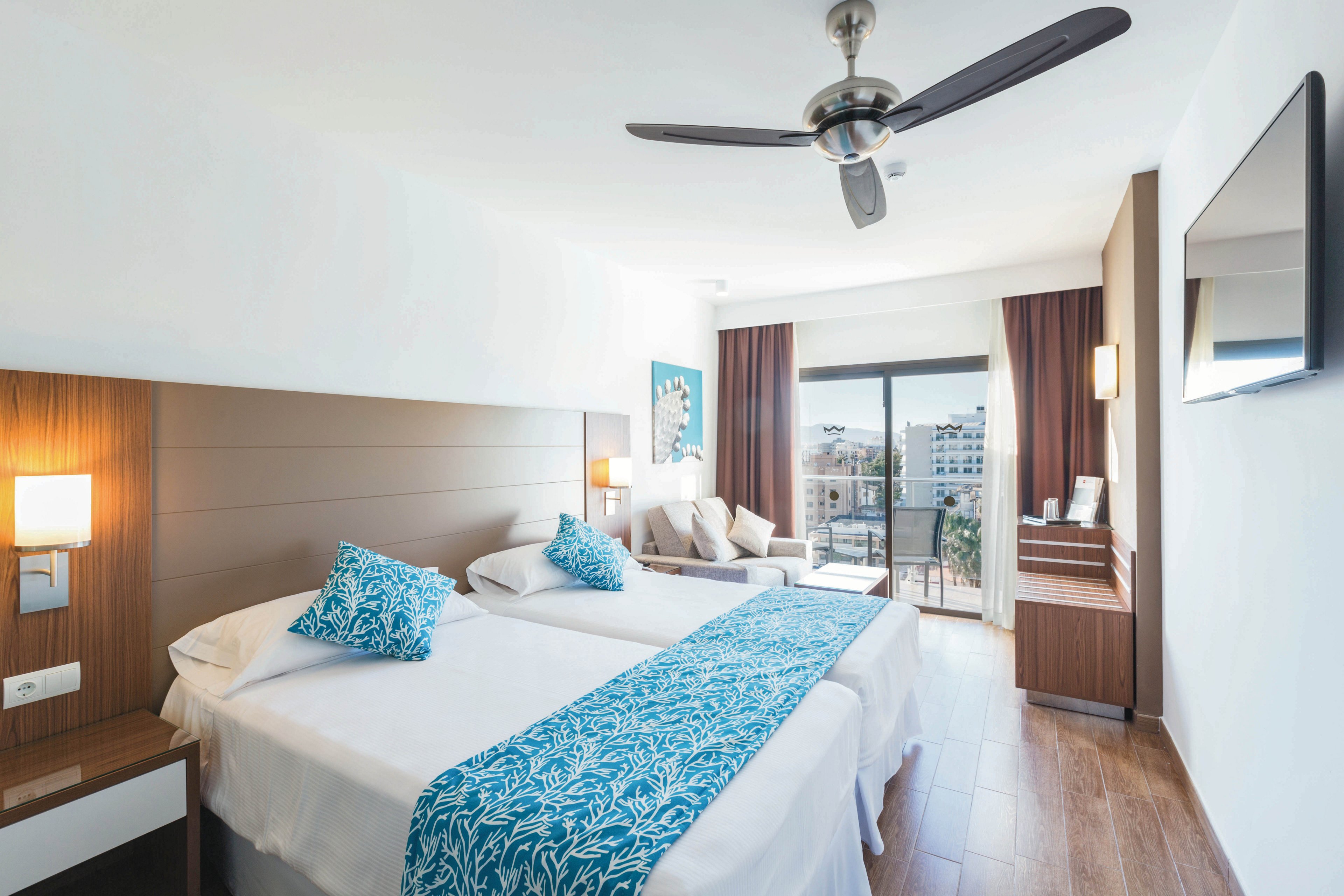 A bright, contemporary hotel room features two twin beds with white linens and blue patterned accents. The space includes wooden furniture, a wall-mounted TV, and a ceiling fan, with large windows leading to a balcony overlooking a cityscape. Natural light fills the room, highlighting the clean, minimalist decor and comfortable seating area. The overall mood is fresh and inviting, ideal for relaxation or business travel.