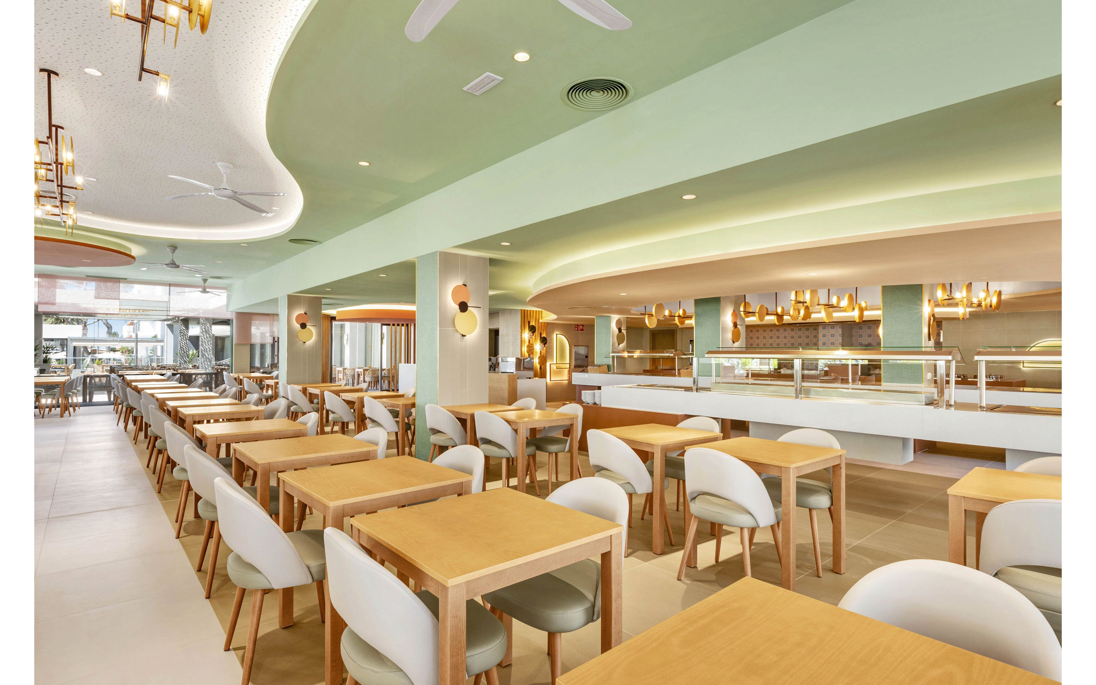 Spacious restaurant dining area featuring light wood tables and white cushioned chairs arranged in neat rows. The setting is bright and airy, with pastel green accents and contemporary lighting fixtures. The open buffet counter and ceiling fans add to the modern, inviting atmosphere. No visible people or text are present in the image.