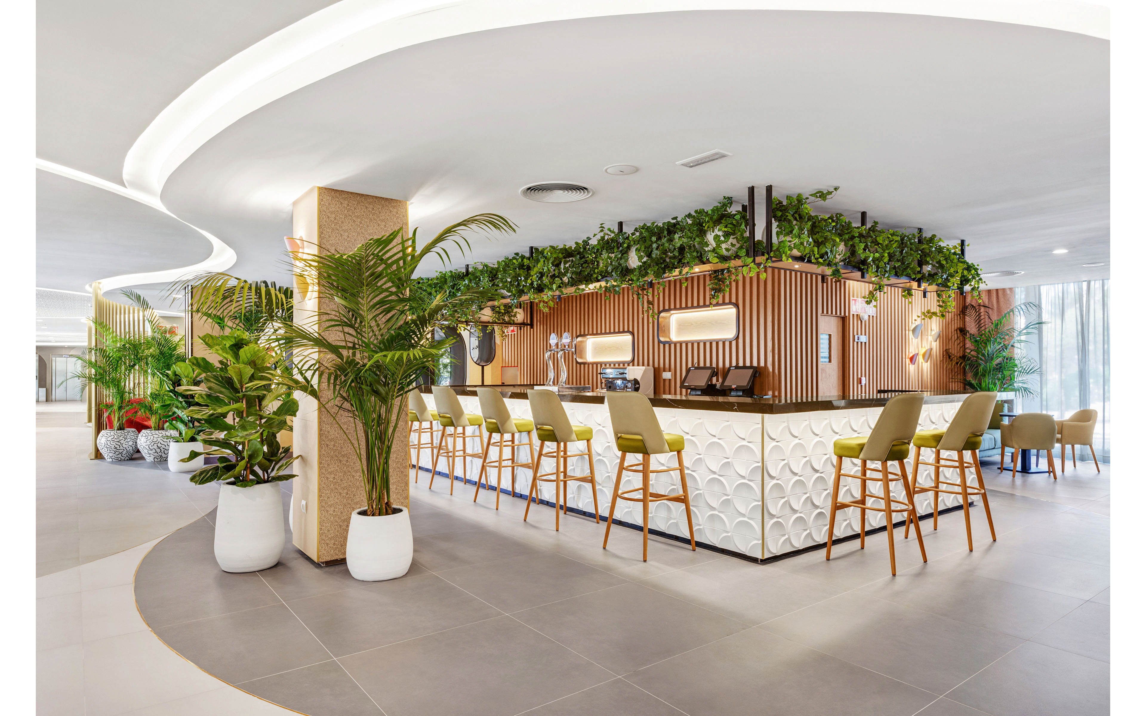 A contemporary hotel bar features a geometric white counter surrounded by green cushioned bar stools. The setting is bright and open, with abundant natural light and lush indoor plants enhancing the fresh ambiance. Overhead, greenery and wood accents add warmth to the modern design. The space is empty, emphasizing the clean lines and inviting atmosphere. No visible text or numbers are present in the image.