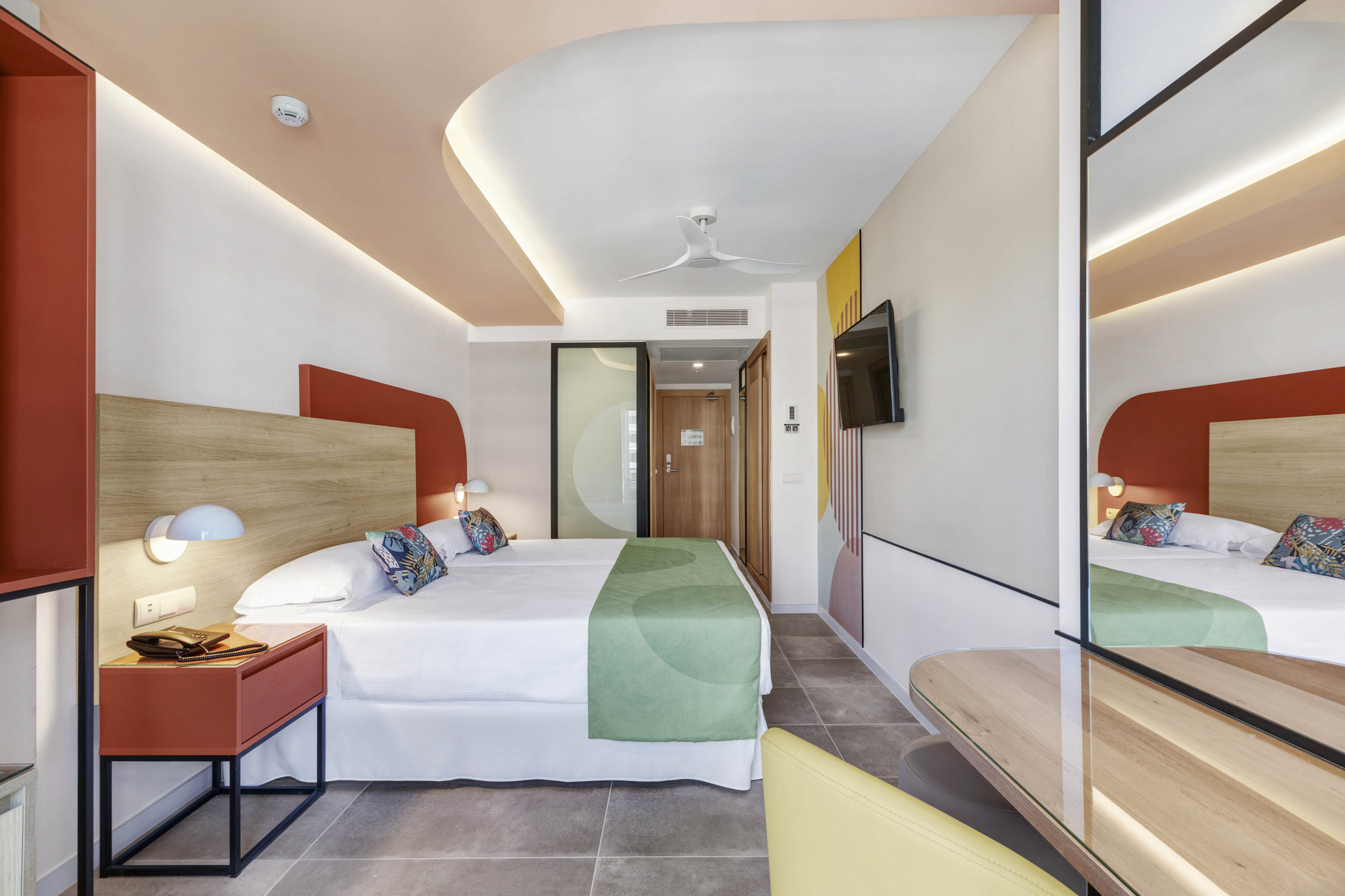 A contemporary hotel room features a double bed with a green runner and colorful accent pillows. The setting includes a wall-mounted television, bedside tables, and a large mirror reflecting the space. The room is well-lit with a neutral palette accented by pops of red and green, and the overall atmosphere is clean and inviting. No visible text or numbers are present in the image.