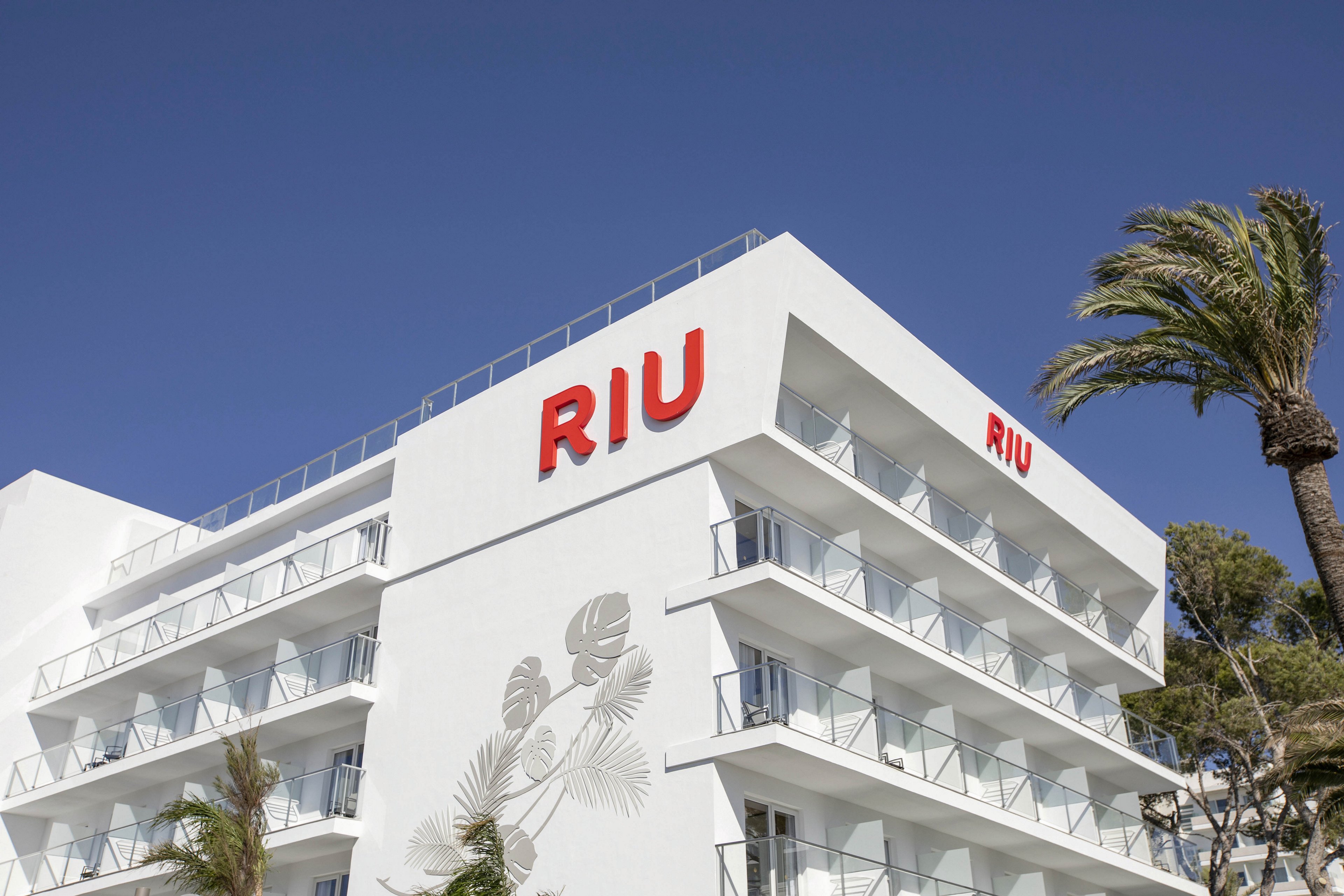 A white multi-story RIU hotel building stands under a clear blue sky. The bold red RIU signage is prominently displayed on the facade, with balconies and glass railings visible. Palm trees and subtle wall art add a tropical atmosphere to the scene. The overall mood is bright and inviting, suggesting a vacation or resort setting.
