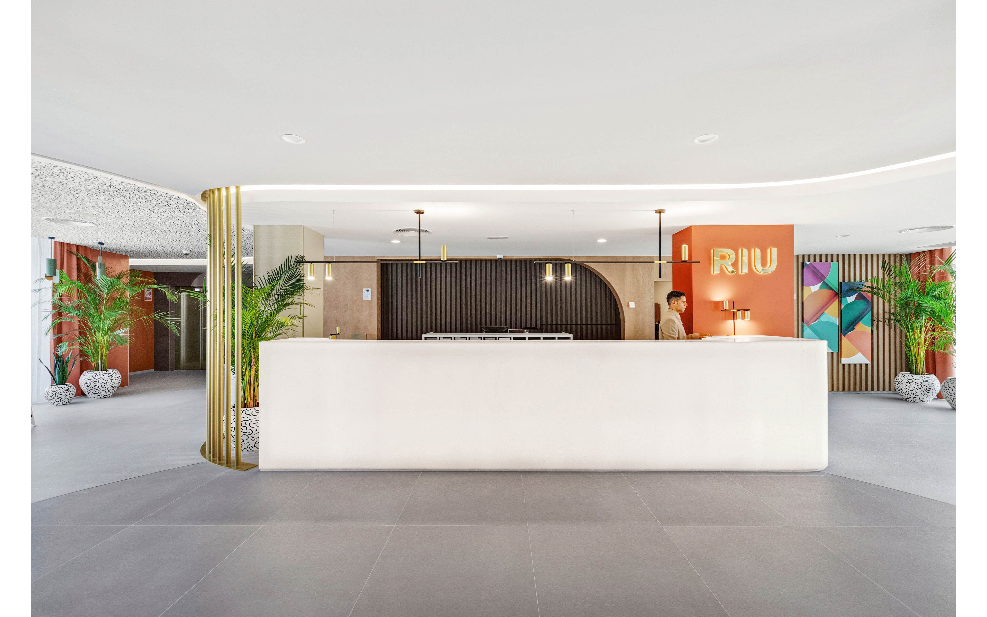 A spacious, contemporary hotel reception area features a large white desk and a staff member standing behind it. The setting is bright and minimalist, with decorative plants and geometric wall art adding visual interest. The word 'RIU' is clearly visible on an orange wall panel. Warm lighting and neutral tones create a welcoming atmosphere.
