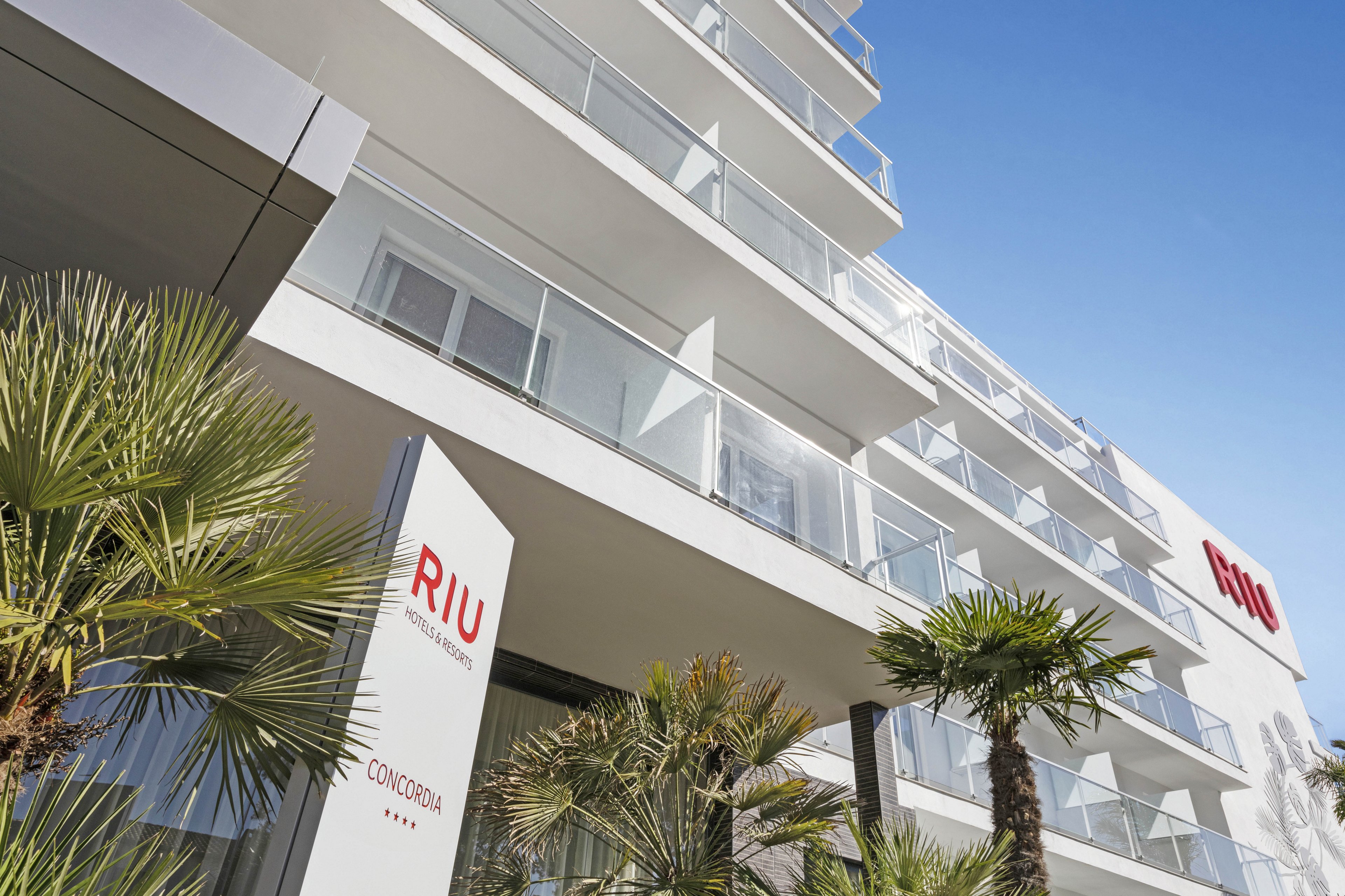 The image shows the exterior of the RIU Concordia hotel, featuring a modern white facade with glass balconies. Palm trees are visible in the foreground, adding a tropical atmosphere to the scene. The sky is clear and blue, enhancing the bright and inviting mood. The hotel name 'RIU' and 'CONCORDIA' are clearly visible on signage, along with four stars.