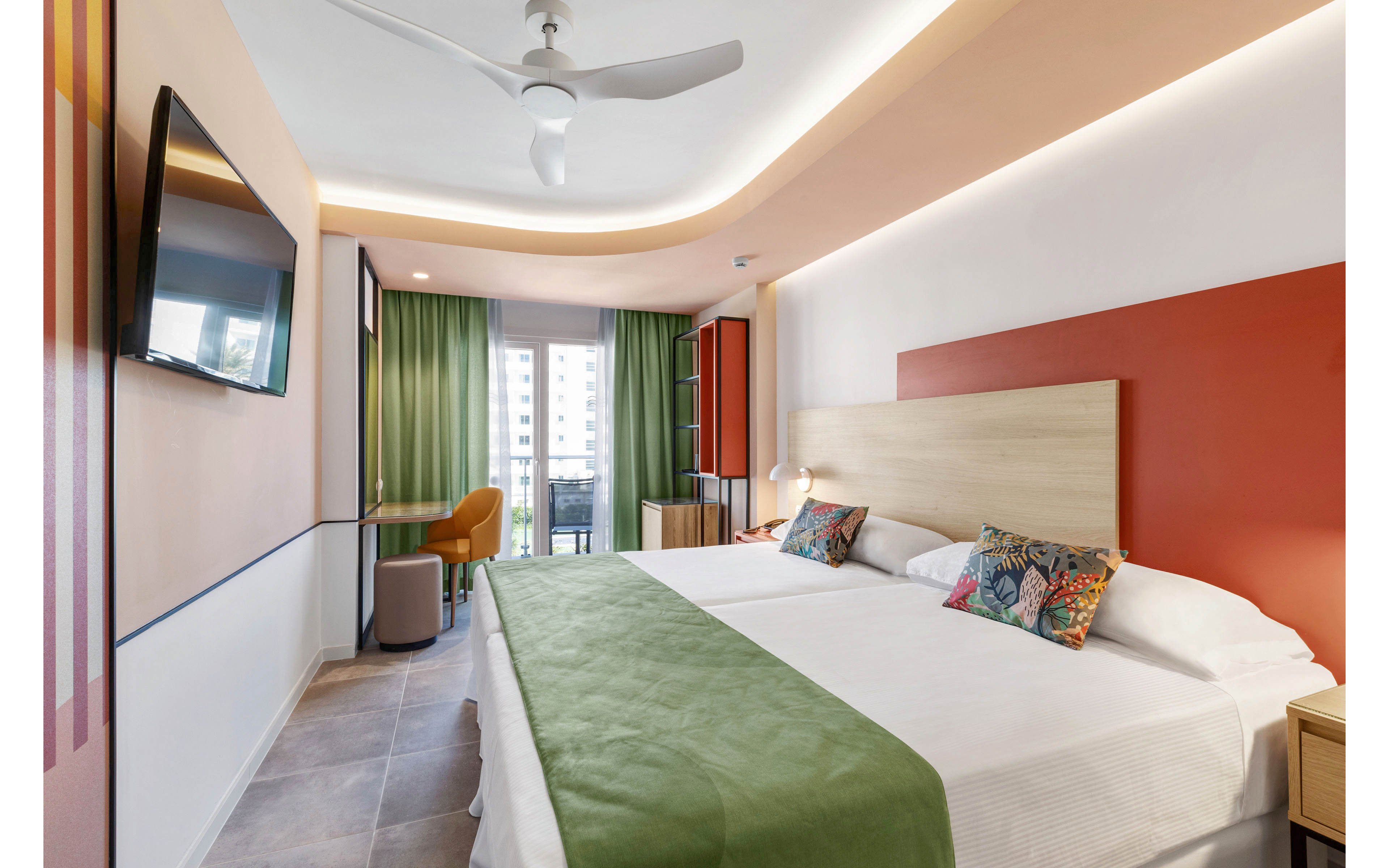 A contemporary hotel room features two twin beds with green runners and colorful pillows. The space is well-lit, with a ceiling fan, wall-mounted TV, and a work desk near the window. Large glass doors open to a balcony with city views, and the room is decorated in warm tones with minimalist furnishings. No visible text or numbers are present in the image.