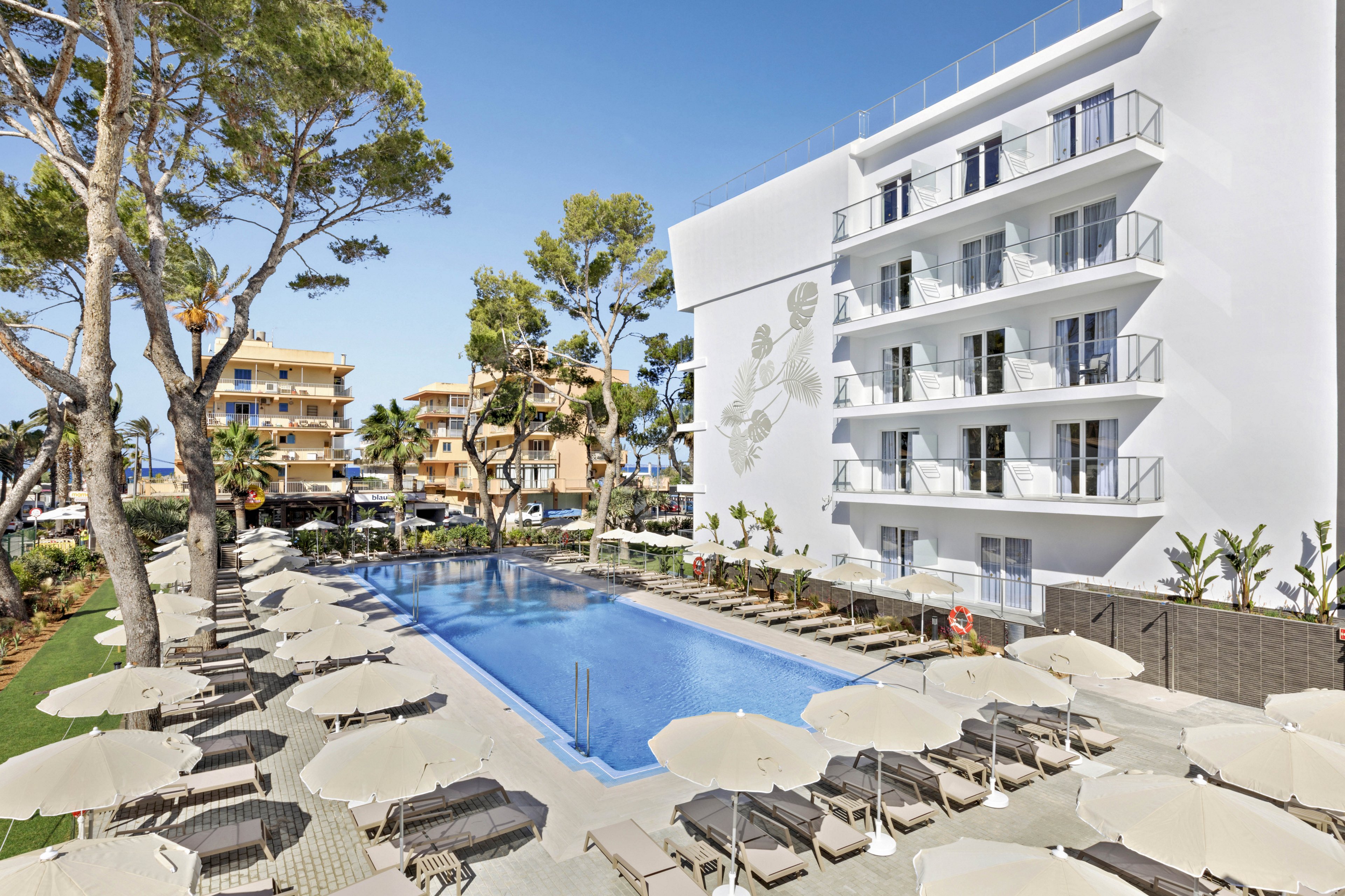 A contemporary hotel building features multiple balconies overlooking a rectangular outdoor swimming pool. Numerous sun loungers and umbrellas are arranged around the pool, creating a relaxing atmosphere. Tall trees and nearby residential buildings add to the sunny, vacation-like setting. The scene is bright with clear blue skies, emphasizing a tranquil and inviting mood.