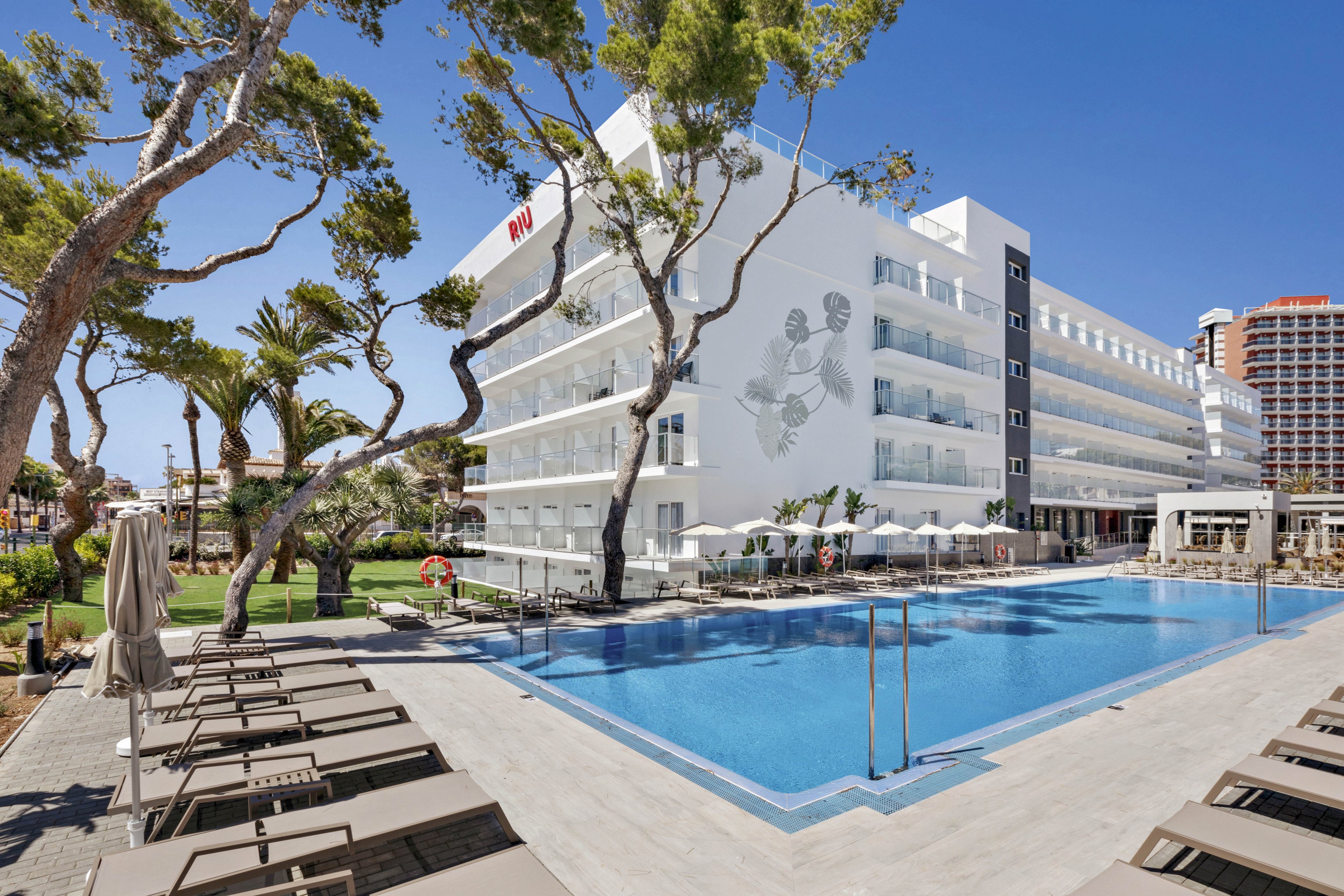 A contemporary white hotel building features a large outdoor swimming pool surrounded by sun loungers and umbrellas. The setting is bright and sunny, with clear blue skies and tall trees providing shade. The hotel facade displays a visible 'RW' text and a mural of athletes. The atmosphere is relaxed, ideal for leisure and vacation stays.