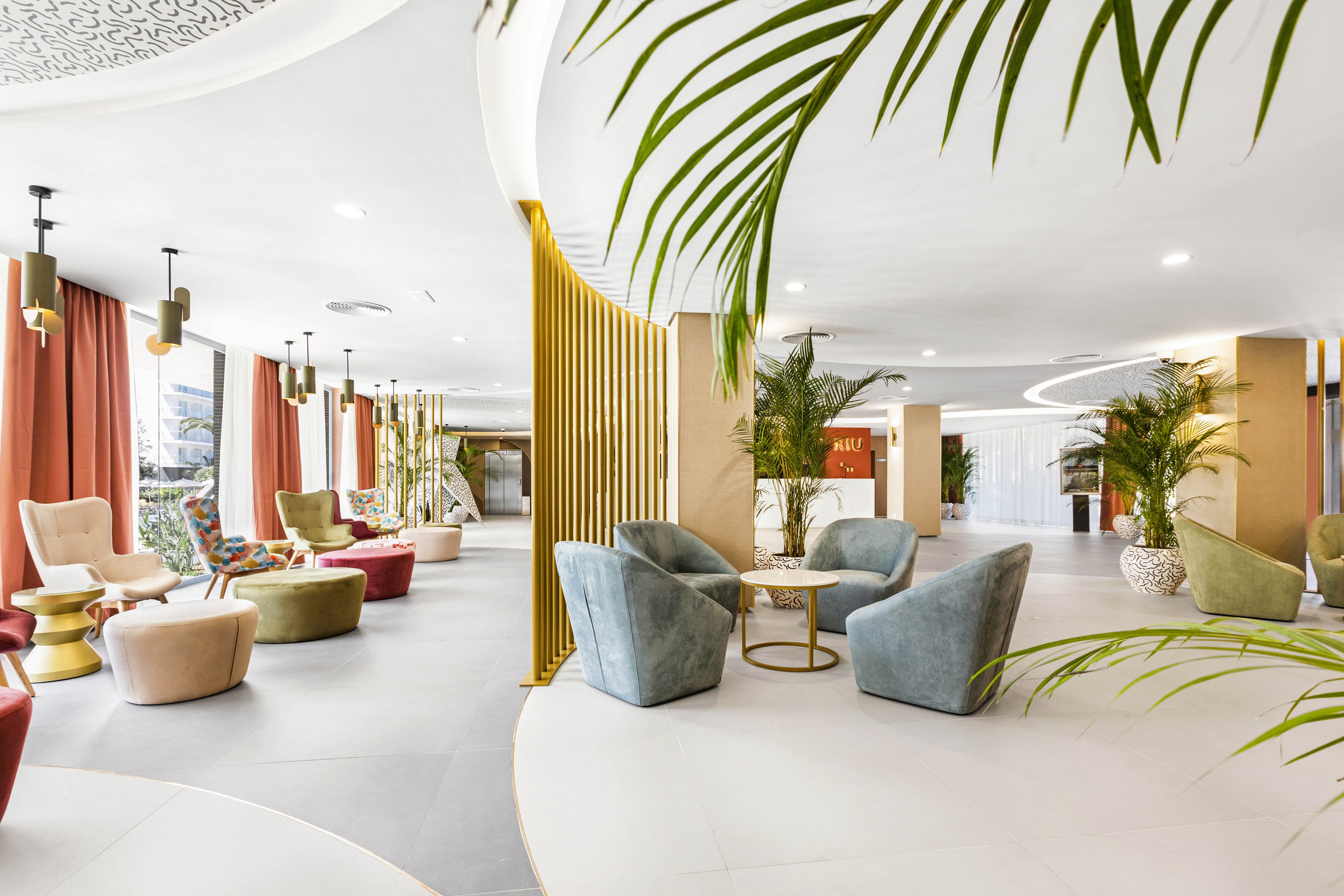 Spacious hotel lobby featuring contemporary armchairs and round tables arranged in small groups. The setting includes large windows, decorative gold dividers, and lush green plants, creating a welcoming and airy atmosphere. Soft pastel tones and natural light enhance the modern, relaxed mood. No visible people or explicit text can be seen in the image.