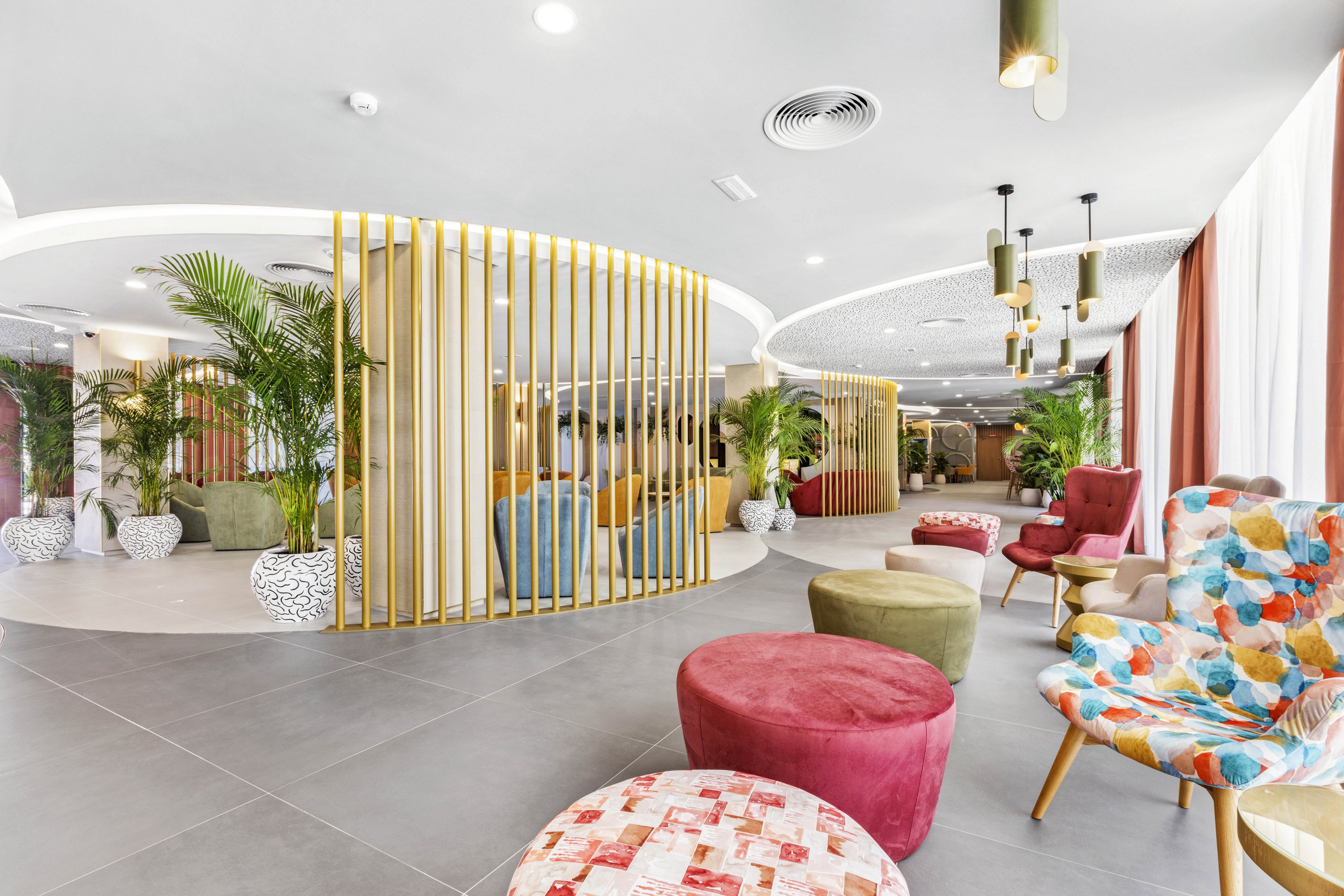 A spacious, contemporary hotel lobby features a variety of colorful chairs and ottomans arranged in a welcoming seating area. Large potted plants and curved wooden dividers add a touch of greenery and separation to the open space. The setting is bright with natural light streaming through large windows, and the decor includes playful patterns and soft pastel tones. The overall mood is inviting and relaxed, ideal for socializing or waiting comfortably.