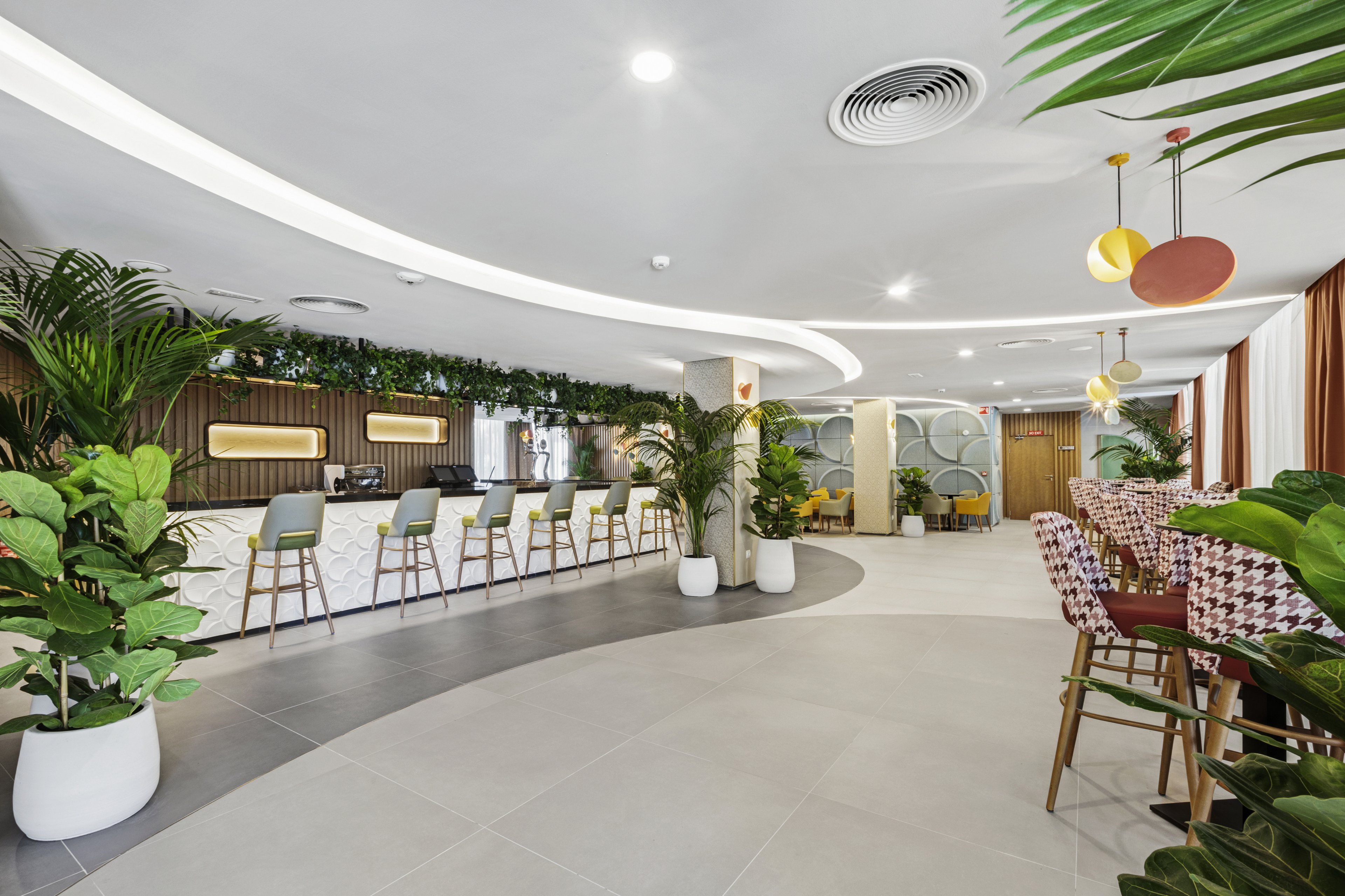 A contemporary hotel lobby features a stylish bar area with high stools and a variety of lush green potted plants. The space is brightly lit with modern ceiling lights and decorated with geometric wall patterns and hanging pendant lamps. The color palette is neutral with pops of green and yellow, creating a welcoming and fresh atmosphere. No visible text or numbers are present in the image.