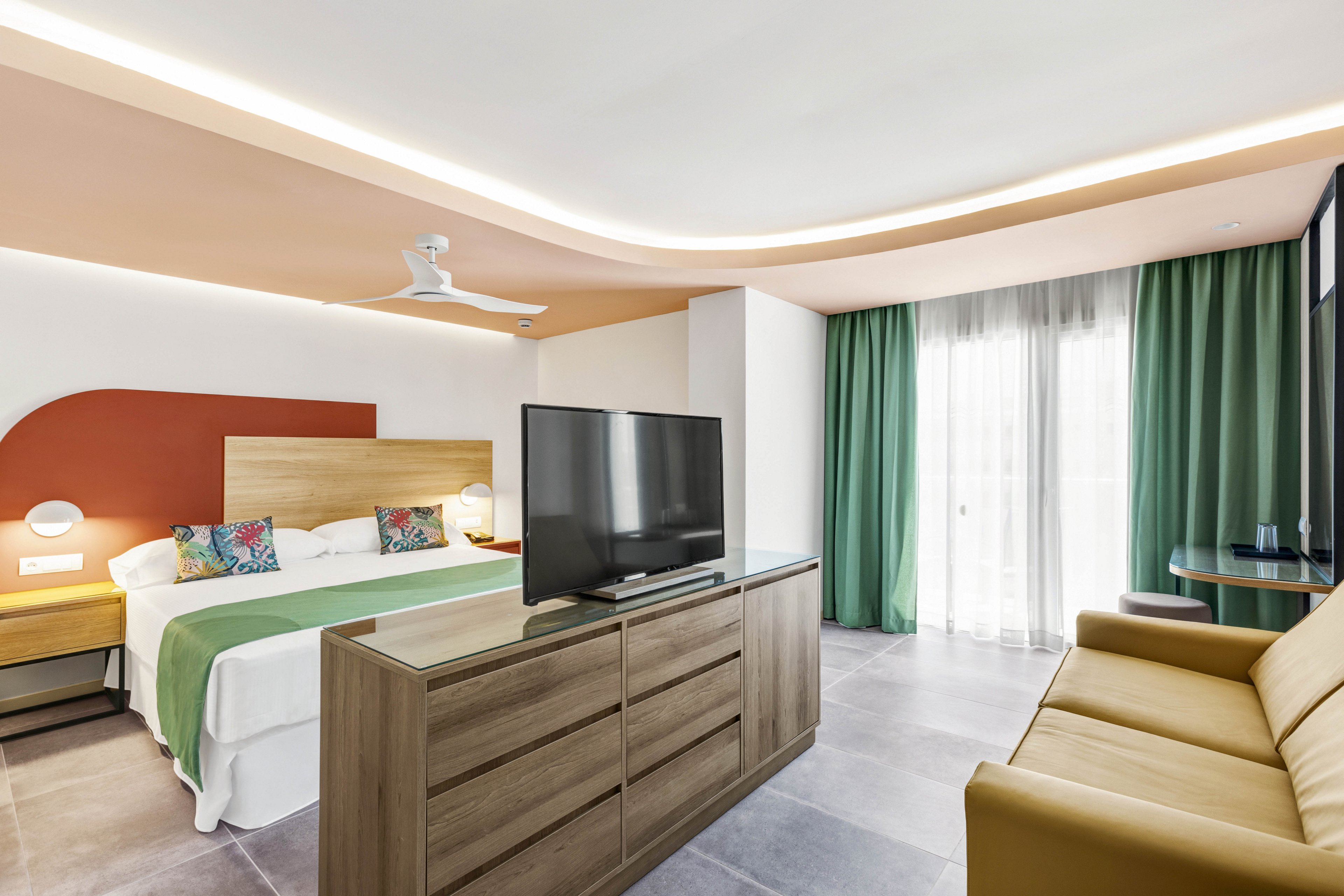 A spacious, contemporary hotel room features a king-sized bed with colorful pillows and a green throw. The room includes a large flat-screen TV on a wooden dresser, a mustard sofa, and a small round table. Natural light enters through sheer curtains, highlighting the minimalist decor and soft, neutral palette.