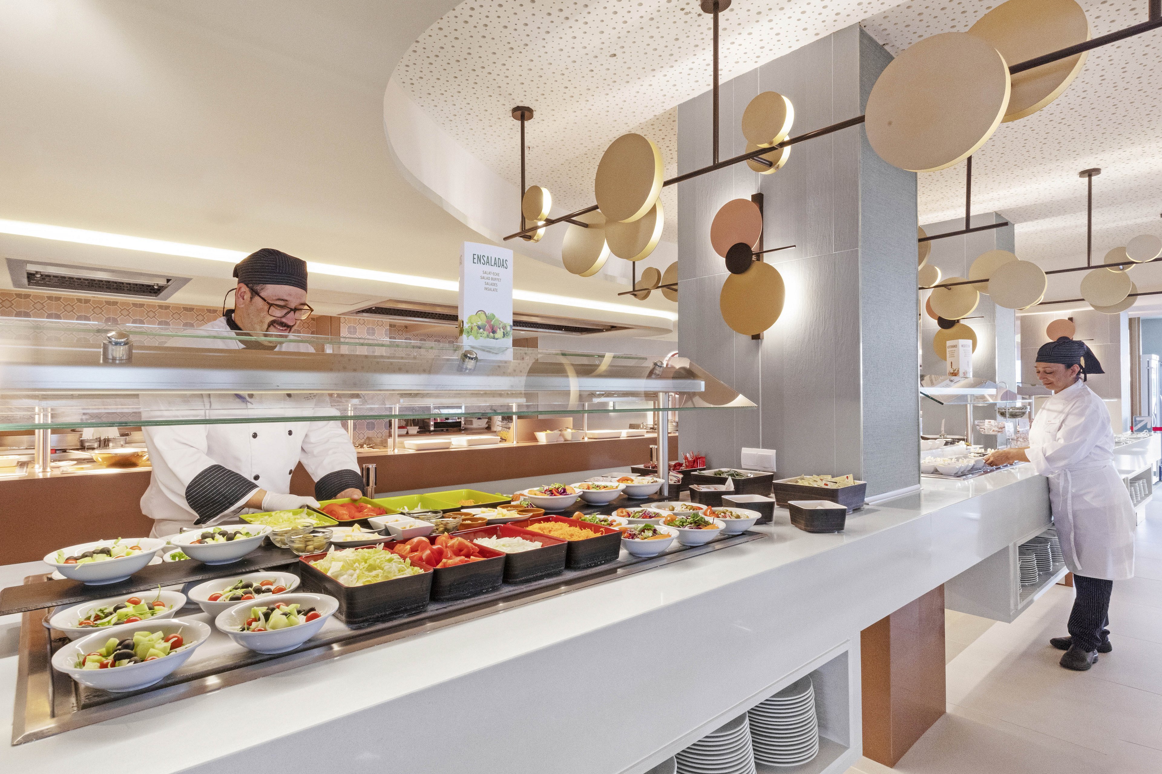 A chef and staff member are working behind a well-lit salad bar in a contemporary cafeteria setting. Various fresh vegetables and salad ingredients are neatly arranged in bowls on the counter. The environment is clean and modern, with geometric decor and bright lighting. Both staff members are dressed in white uniforms and hats, emphasizing hygiene and professionalism. A sign with visible text is placed above the salad bar.