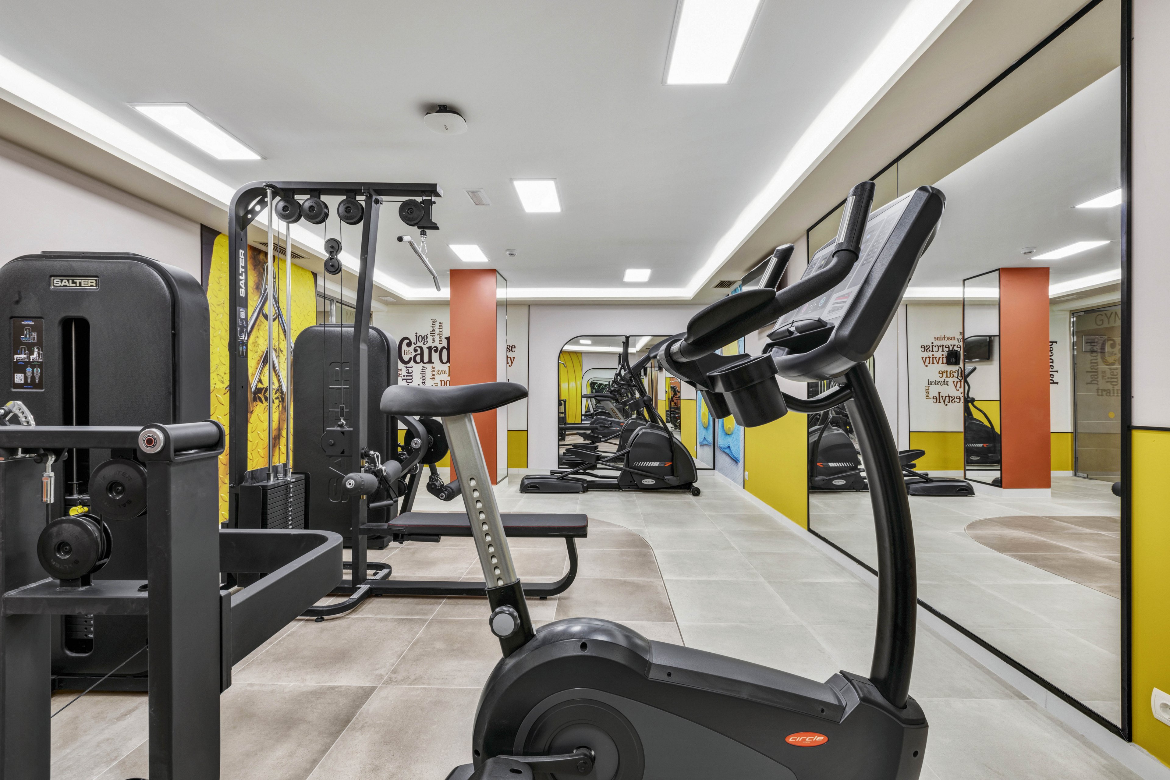 A contemporary gym features a variety of exercise machines, including stationary bikes and strength training equipment. The space is well-lit with overhead lights and has large mirrors along one wall, creating a spacious feel. Yellow and orange accents add energy to the neutral palette, and motivational text is visible on the walls. The gym is empty, emphasizing a clean and organized environment.