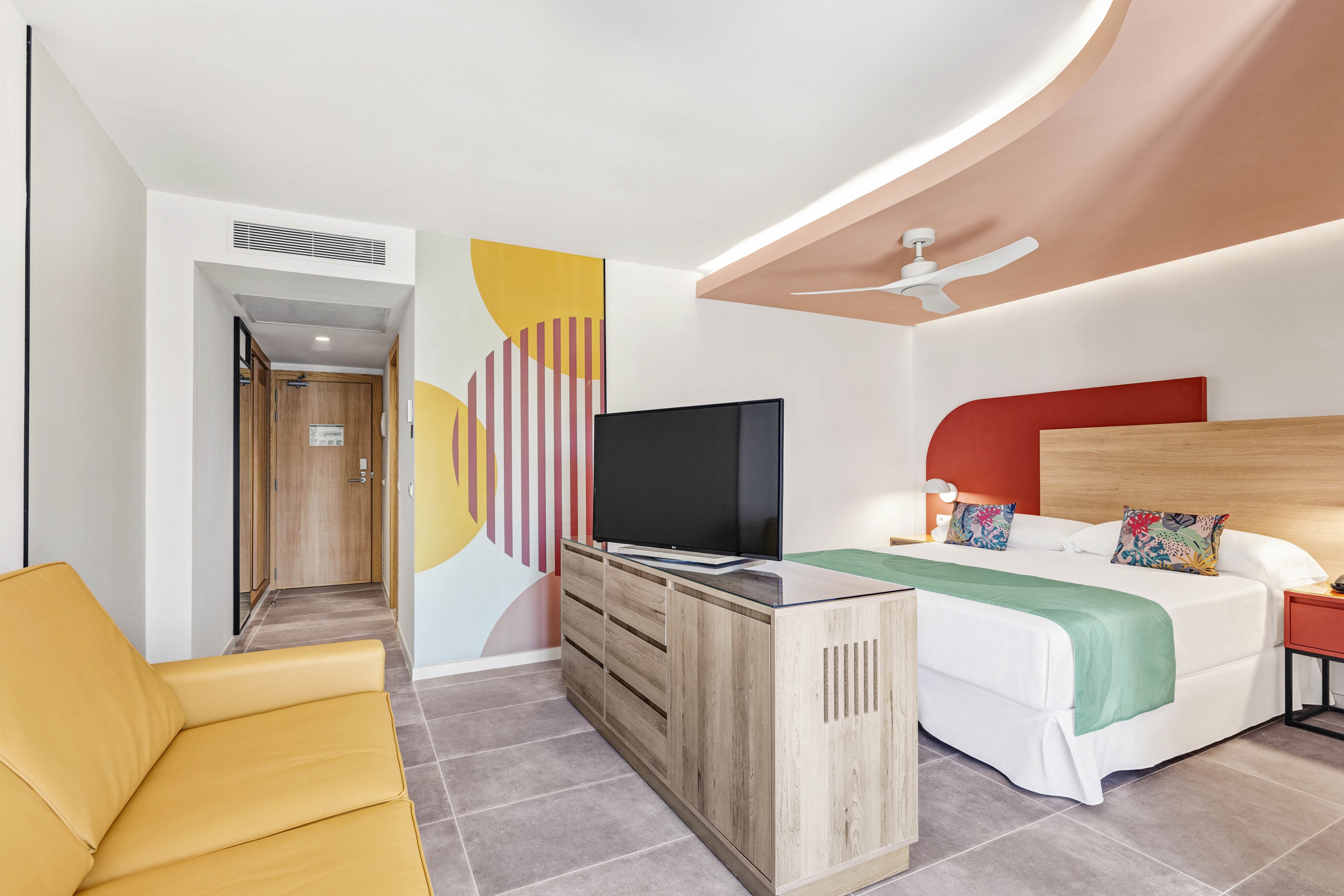 A contemporary hotel room features a king-sized bed with a green runner, vibrant pillows, and a wooden headboard. The space includes a yellow sofa, a large flat-screen TV on a wooden cabinet, and abstract geometric wall art in yellow, red, and purple tones. The room is well-lit, with neutral tile flooring and a ceiling fan, creating a fresh and inviting atmosphere.