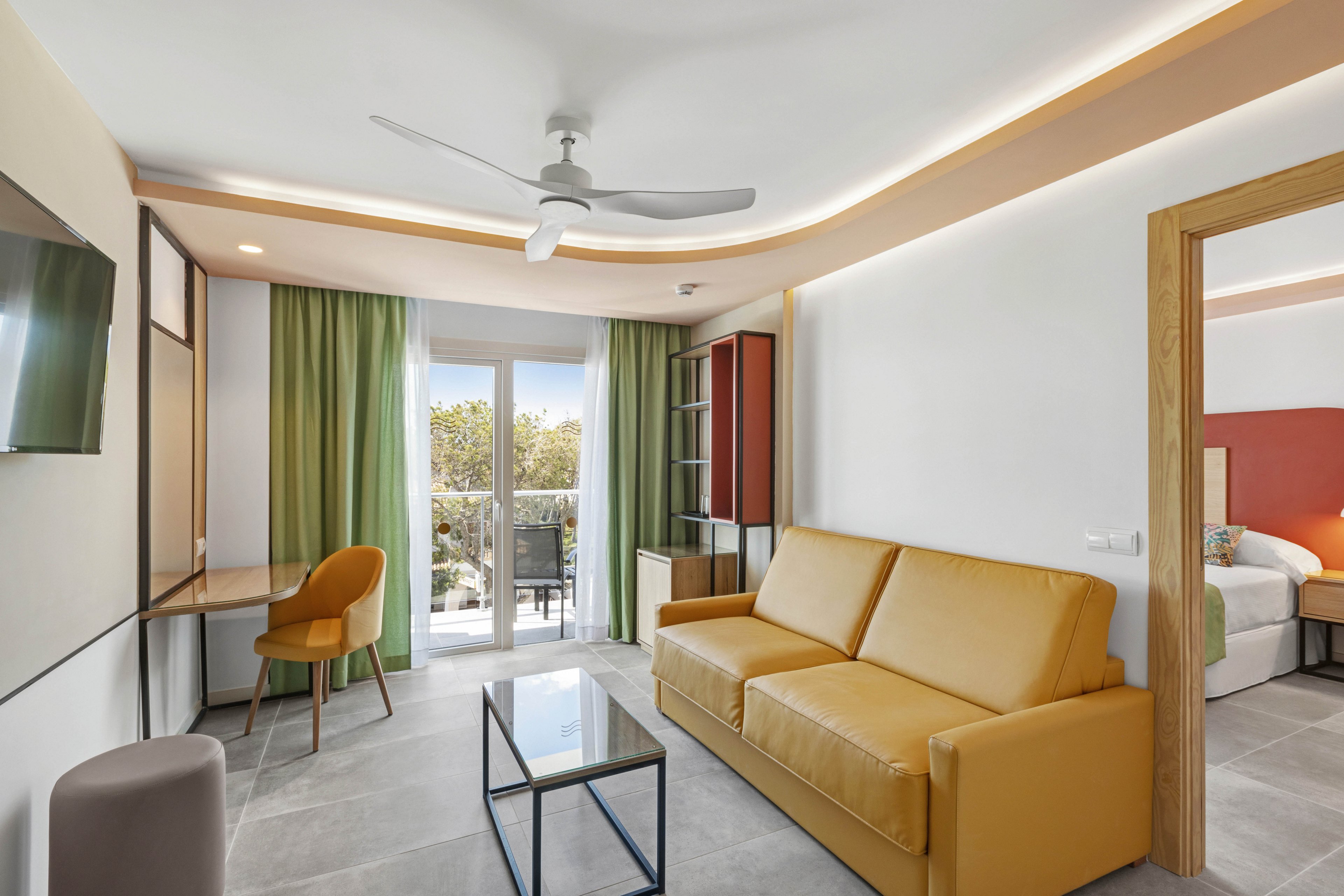 A contemporary hotel suite features a yellow sofa, glass coffee table, and a workspace with a wooden chair. Large windows with green curtains open to a balcony, allowing natural light to fill the room. The adjoining bedroom is partially visible, and the space is accented by neutral tones and minimalist decor. A ceiling fan and wall-mounted TV are also present, enhancing comfort and convenience.