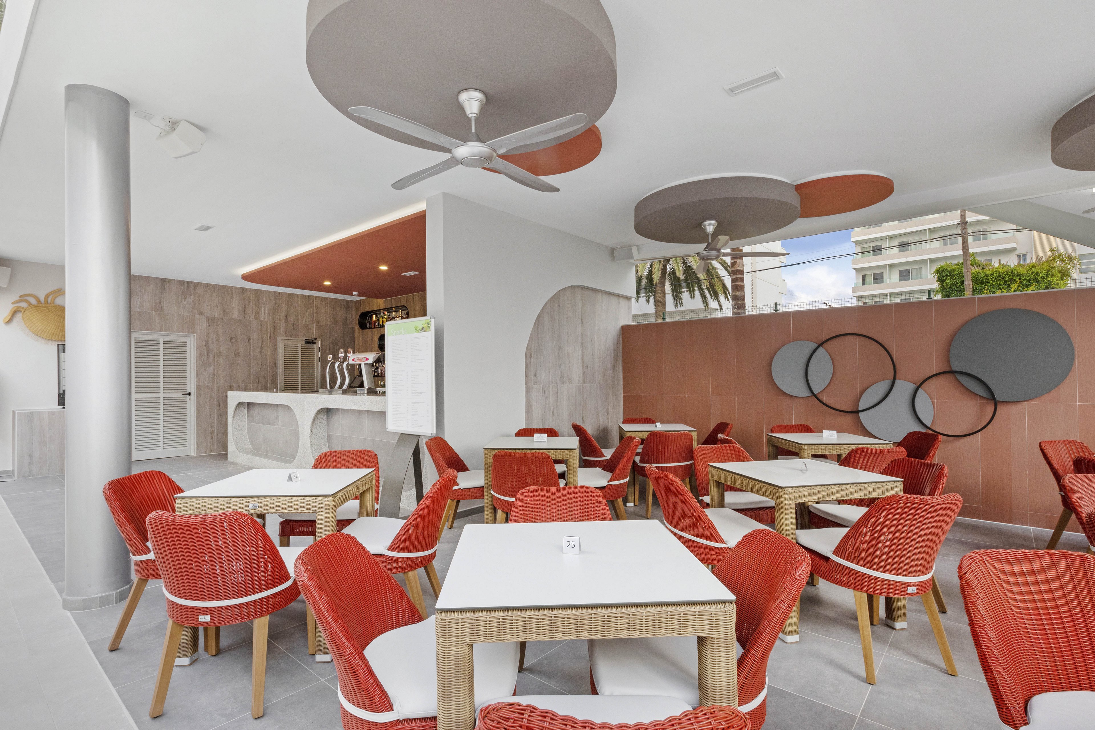 A contemporary cafe features square tables with wicker frames and vibrant red chairs. The setting is bright and airy, with geometric ceiling accents and circular wall decor adding visual interest. Large windows allow natural light to fill the space, and a minimalist bar area is visible in the background. The overall palette is neutral with pops of red, creating a welcoming and stylish atmosphere.