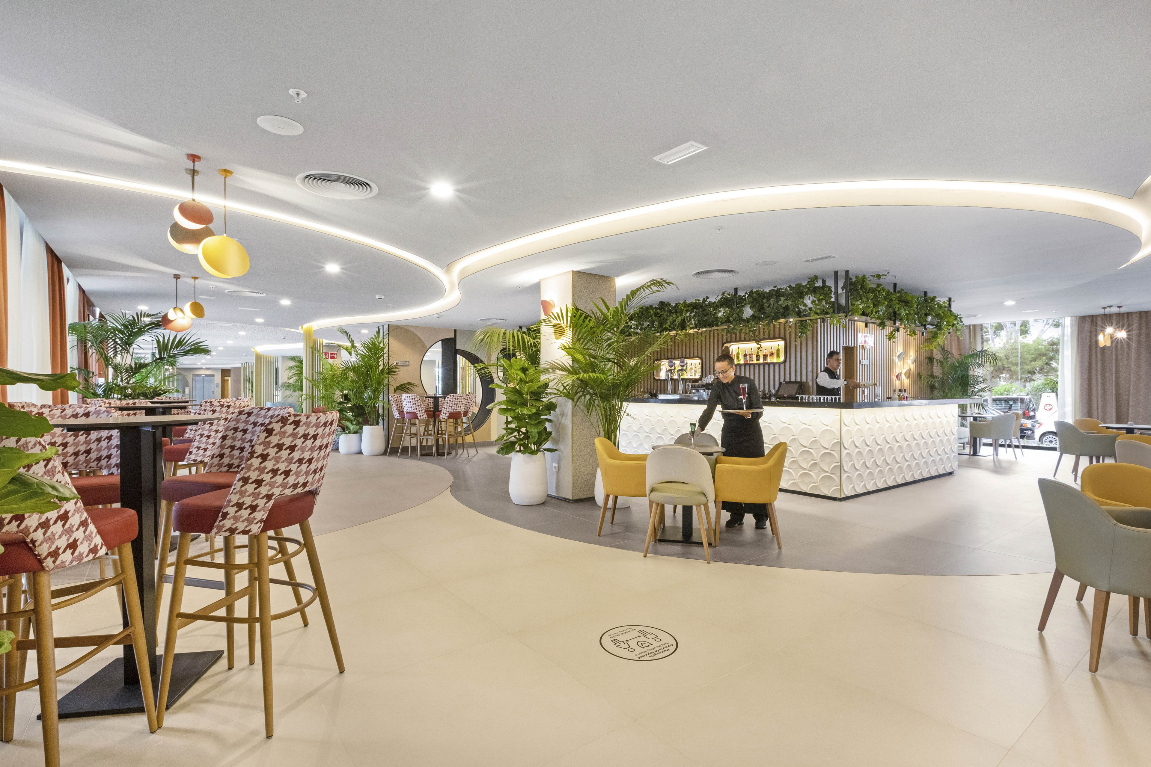 A spacious, contemporary hotel lobby features a stylish bar area with a geometric white counter and hanging greenery. Various seating options, including high stools and cushioned chairs, are arranged throughout the open space. Warm lighting and decorative plants create a welcoming atmosphere, while a person sits at a table near the bar. The overall palette is neutral with pops of color from the furniture.